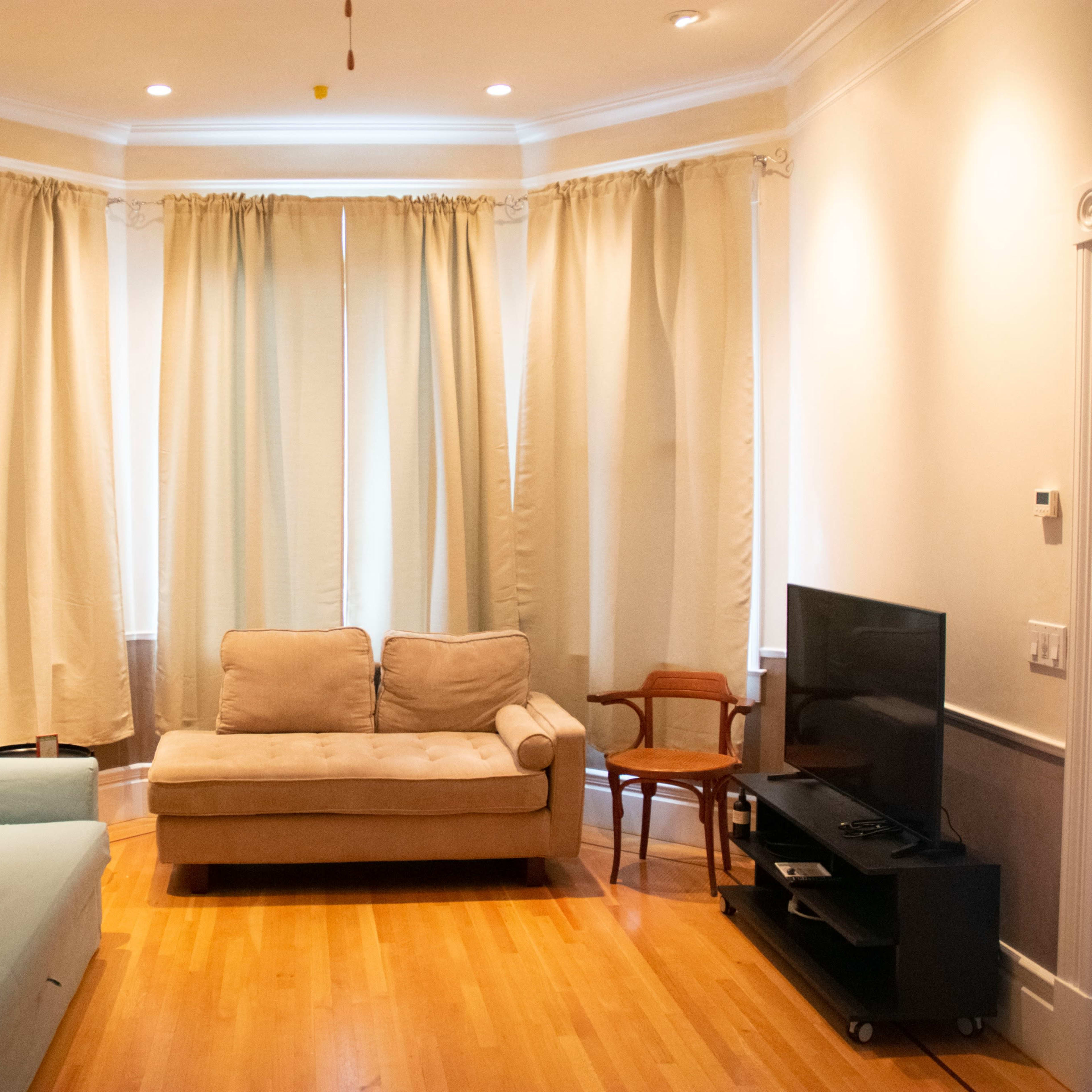A living room with light wooden floors, a beige sofa, a blue couch, a wooden chair, and a television on a black stand, surrounded by light-colored curtains.