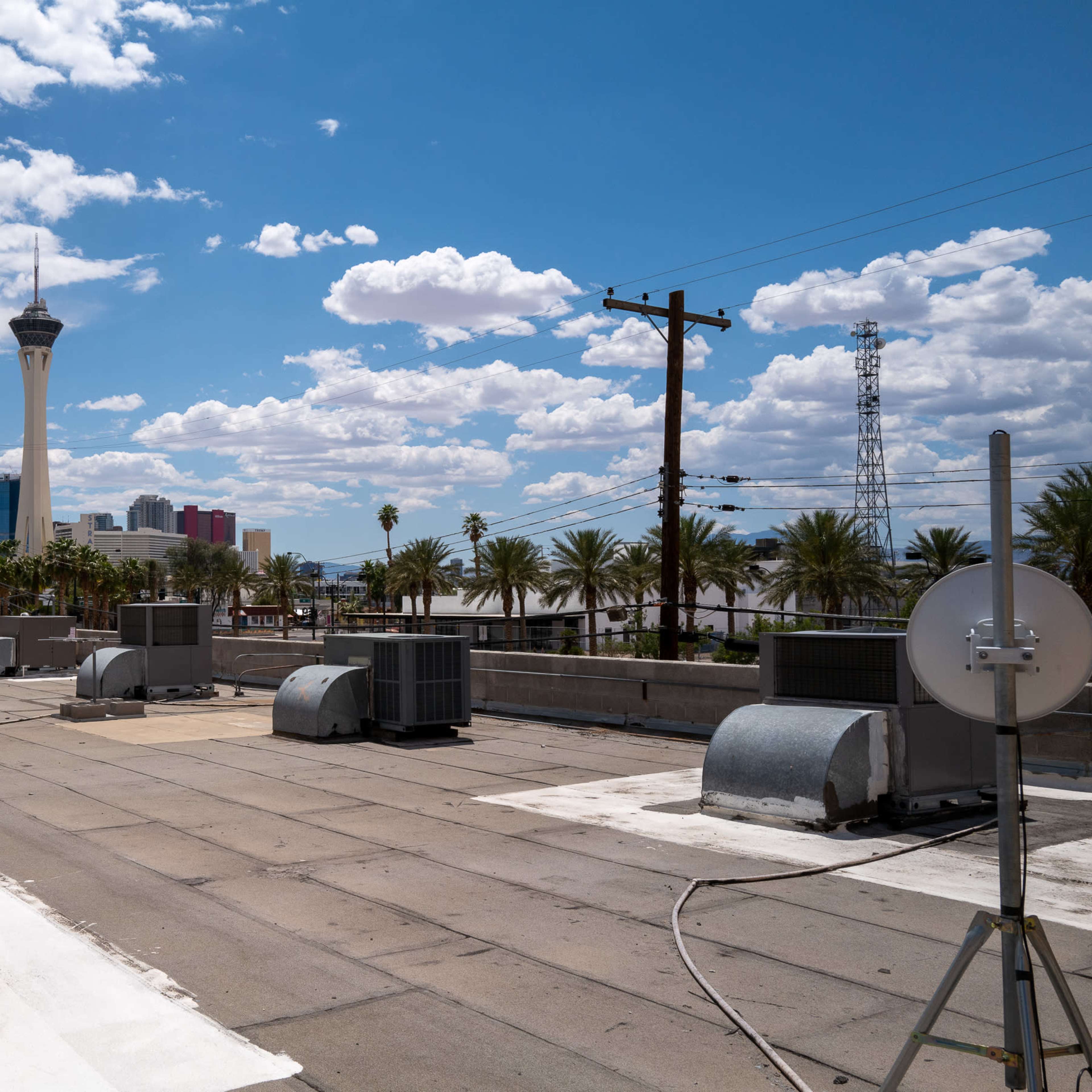 A rooftop view in Las Vegas, featuring air conditioning units, palm trees, and the Stratosphere tower in the background under a partly cloudy sky.