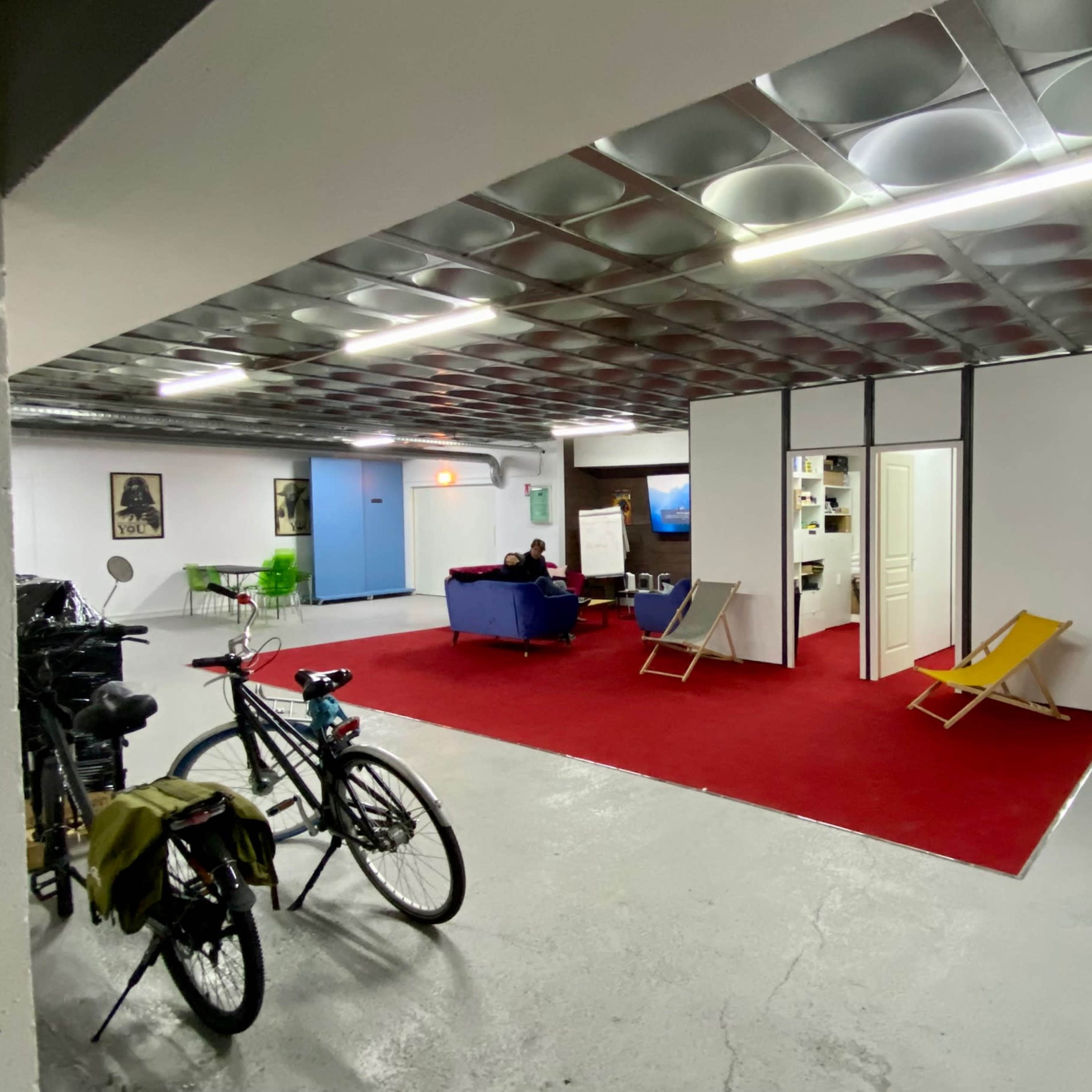 A spacious, multi-functional room with a red carpet, bicycles parked on the left, and seating areas separated by partition walls.