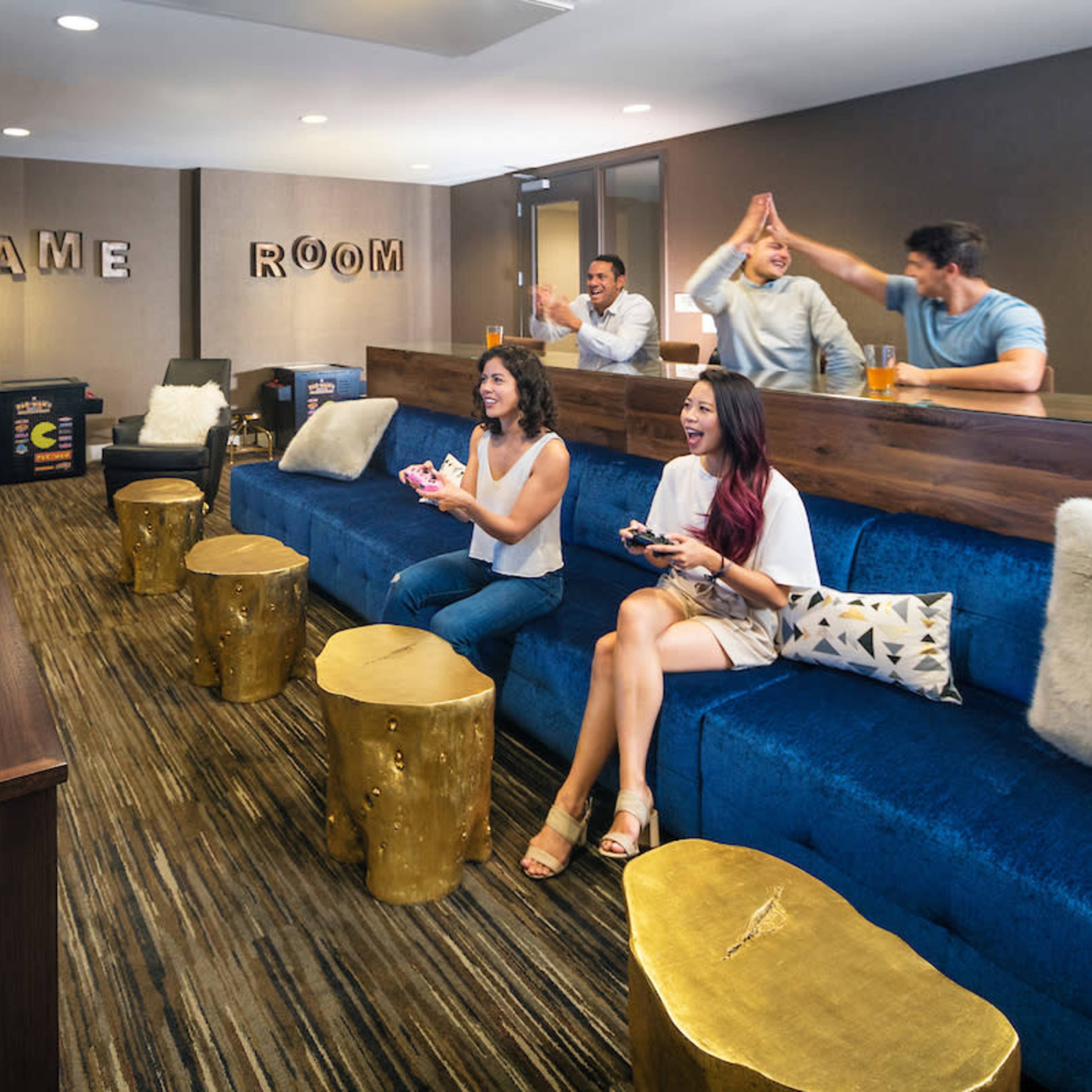 A group of five people is enjoying a gaming session in a modern game room, seated on blue couches with wooden stools in front of a large screen displaying a racing game.