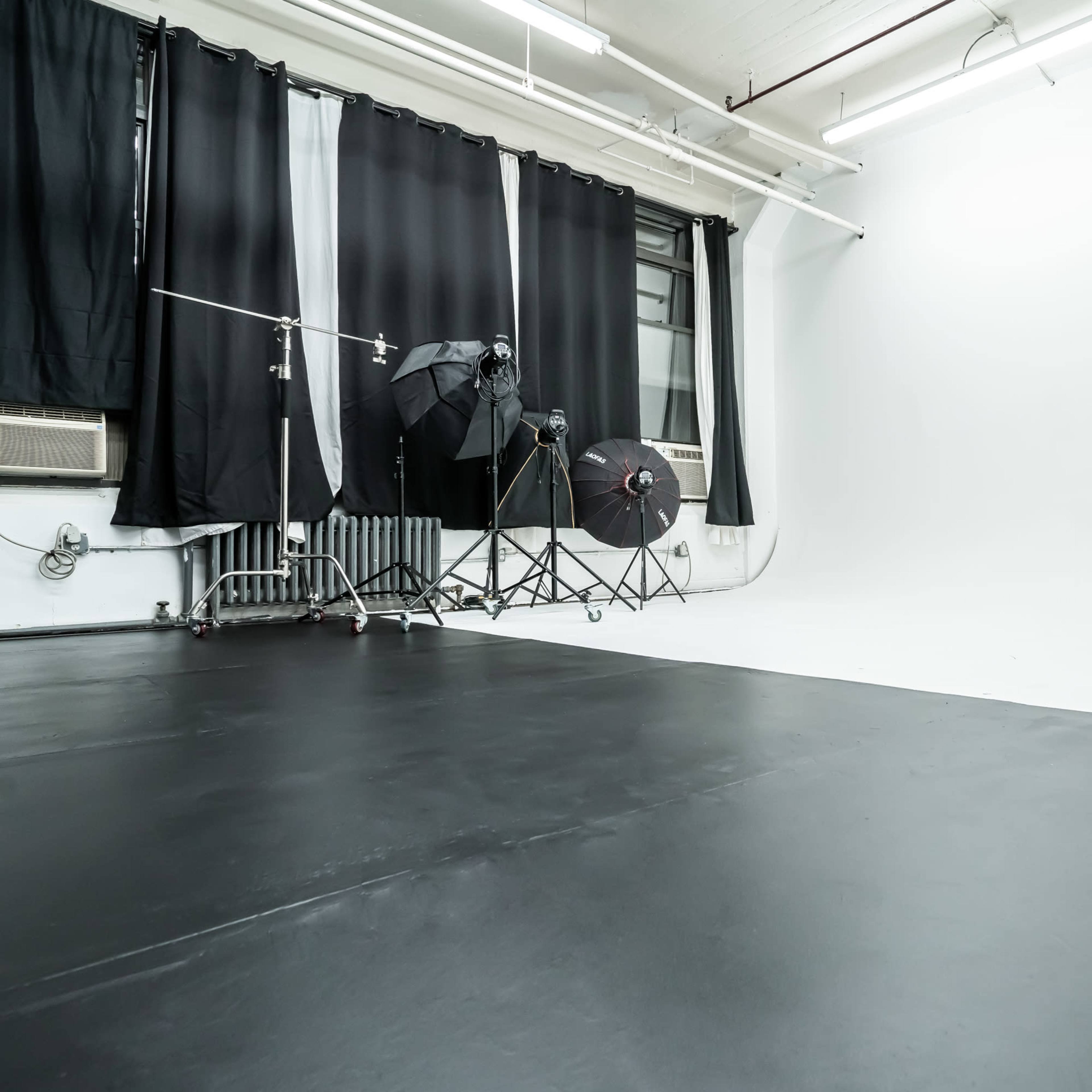 An empty photography studio with black flooring, backdrop curtains, and lighting equipment positioned in the corner.