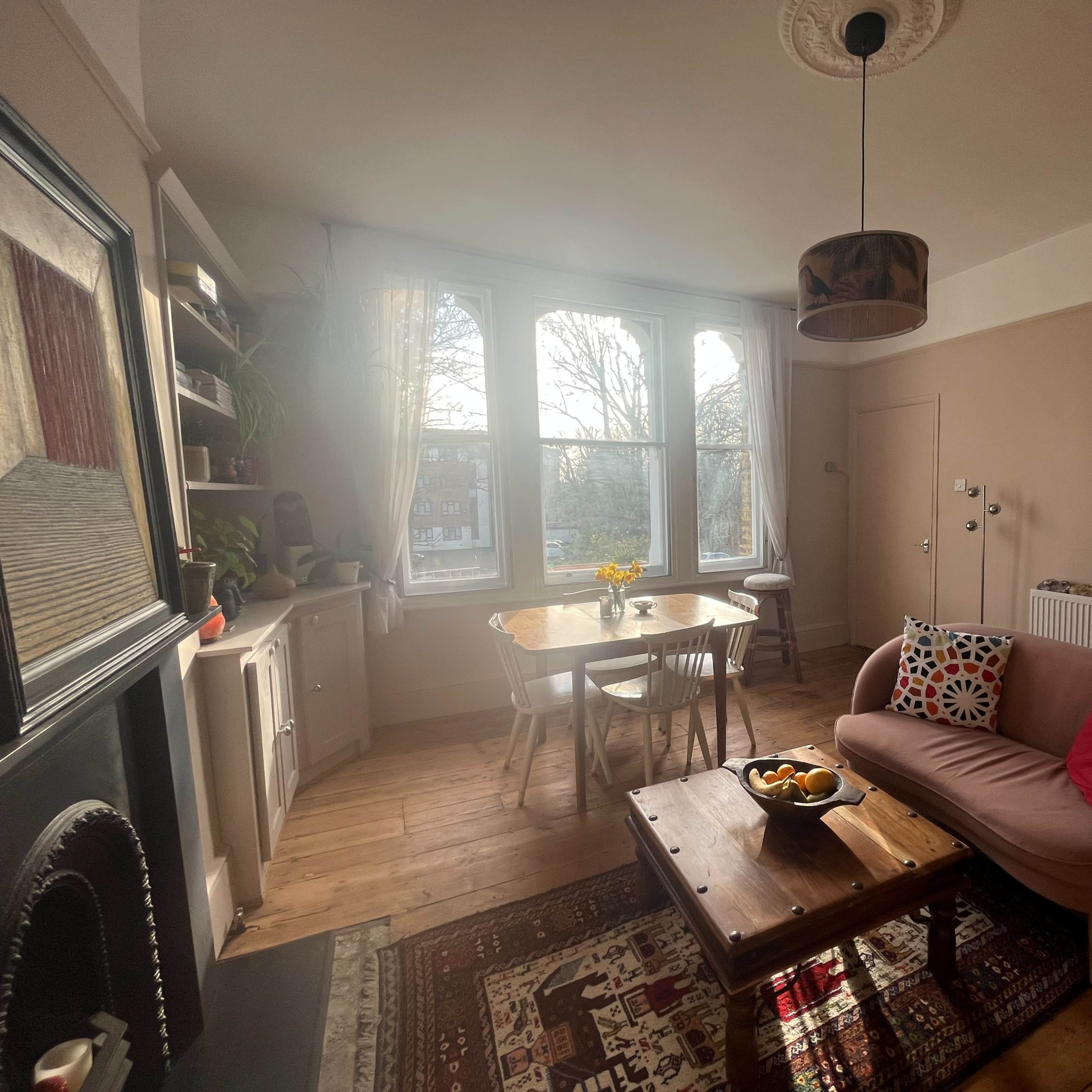 A cozy living room features a wooden floor, a round table with chairs, a sofa adorned with cushions, and large windows letting in natural light.