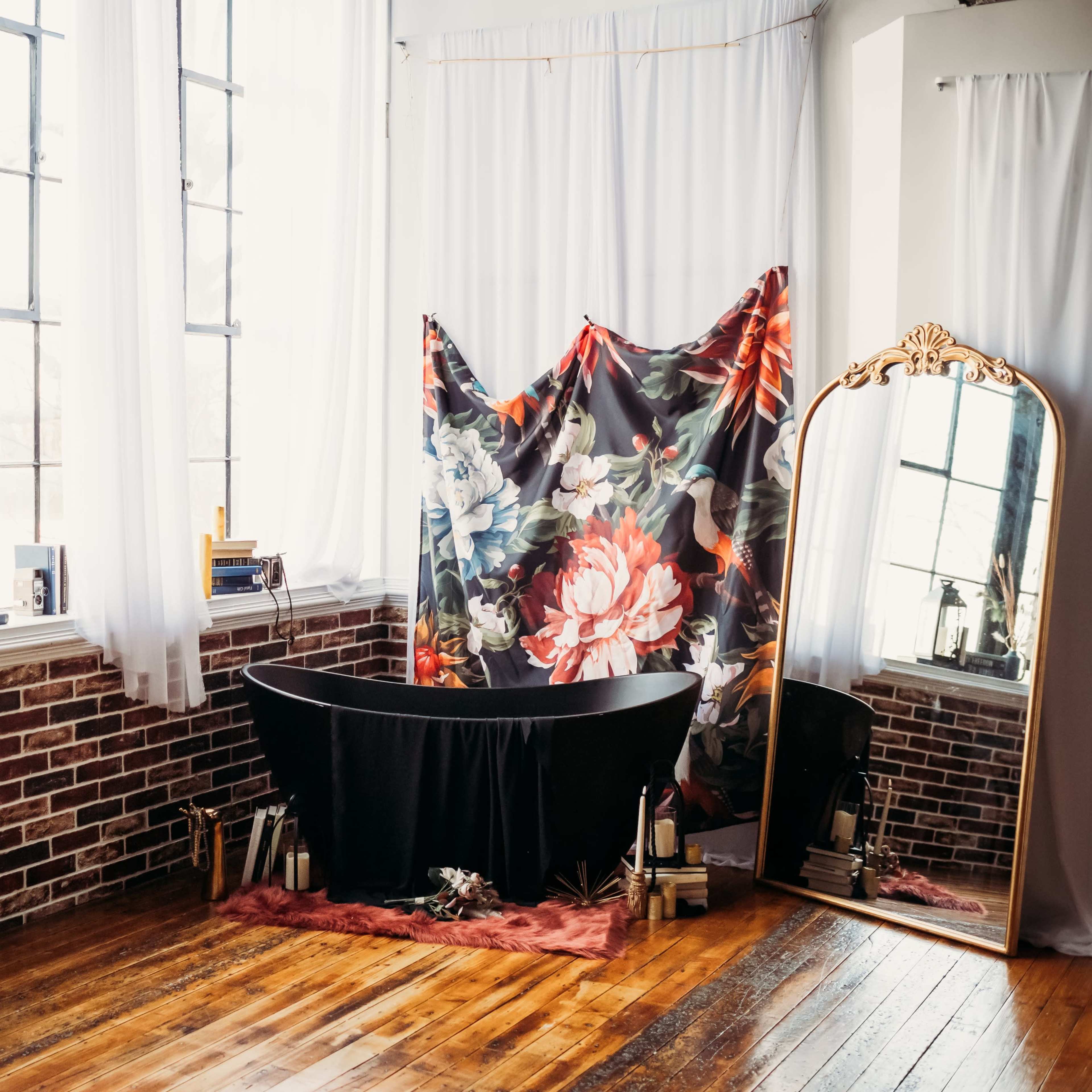 A minimalistic room with large windows, a freestanding black bathtub, a floral backdrop, a mirror, and wooden flooring.