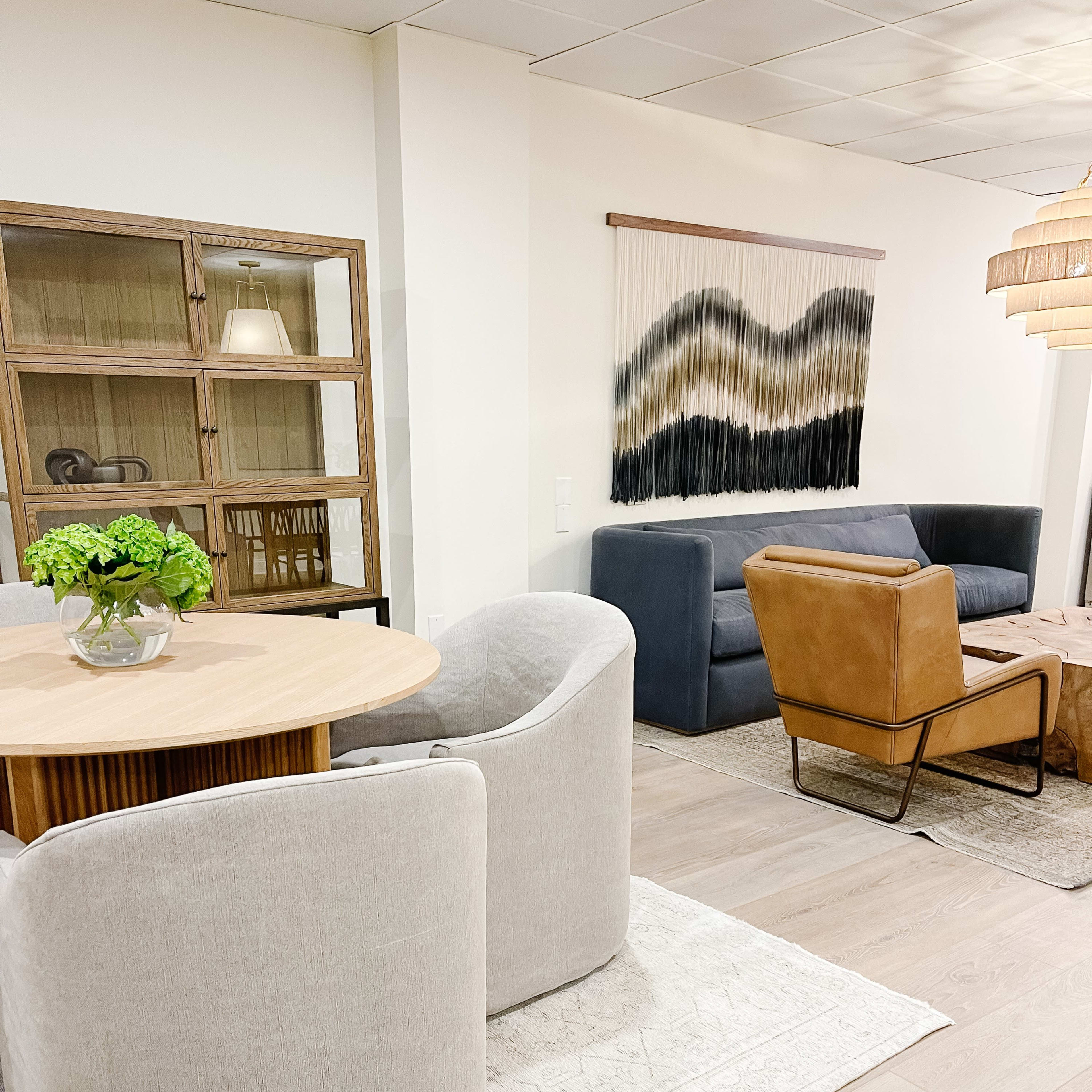 A modern living space featuring a round dining table with curved chairs, a wooden display cabinet, a textured wall hanging, and a mix of seating arrangements including a leather armchair and a gray sofa.