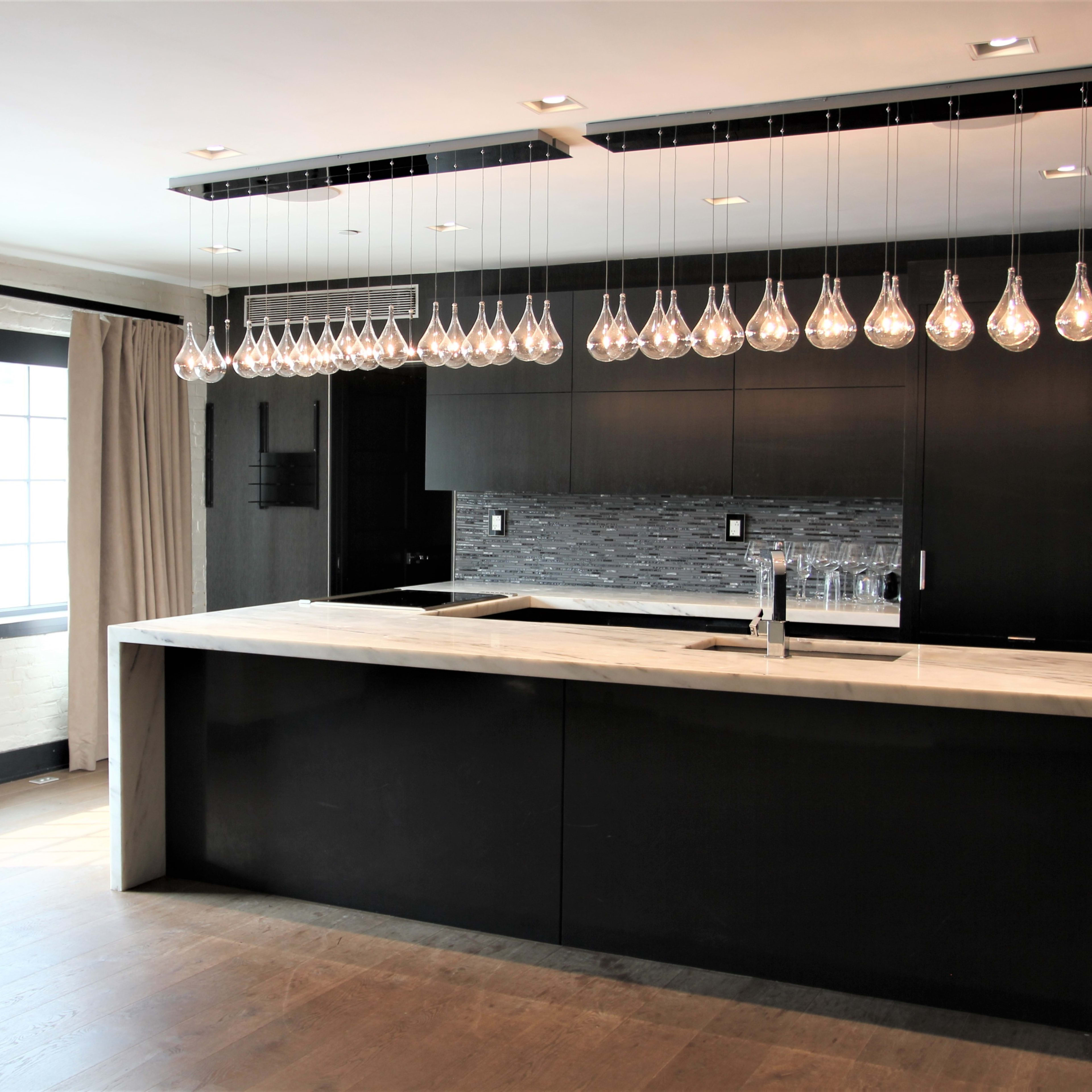 A modern kitchen featuring a large island with a marble countertop, dark cabinetry, and a row of glass pendant lights hanging overhead.