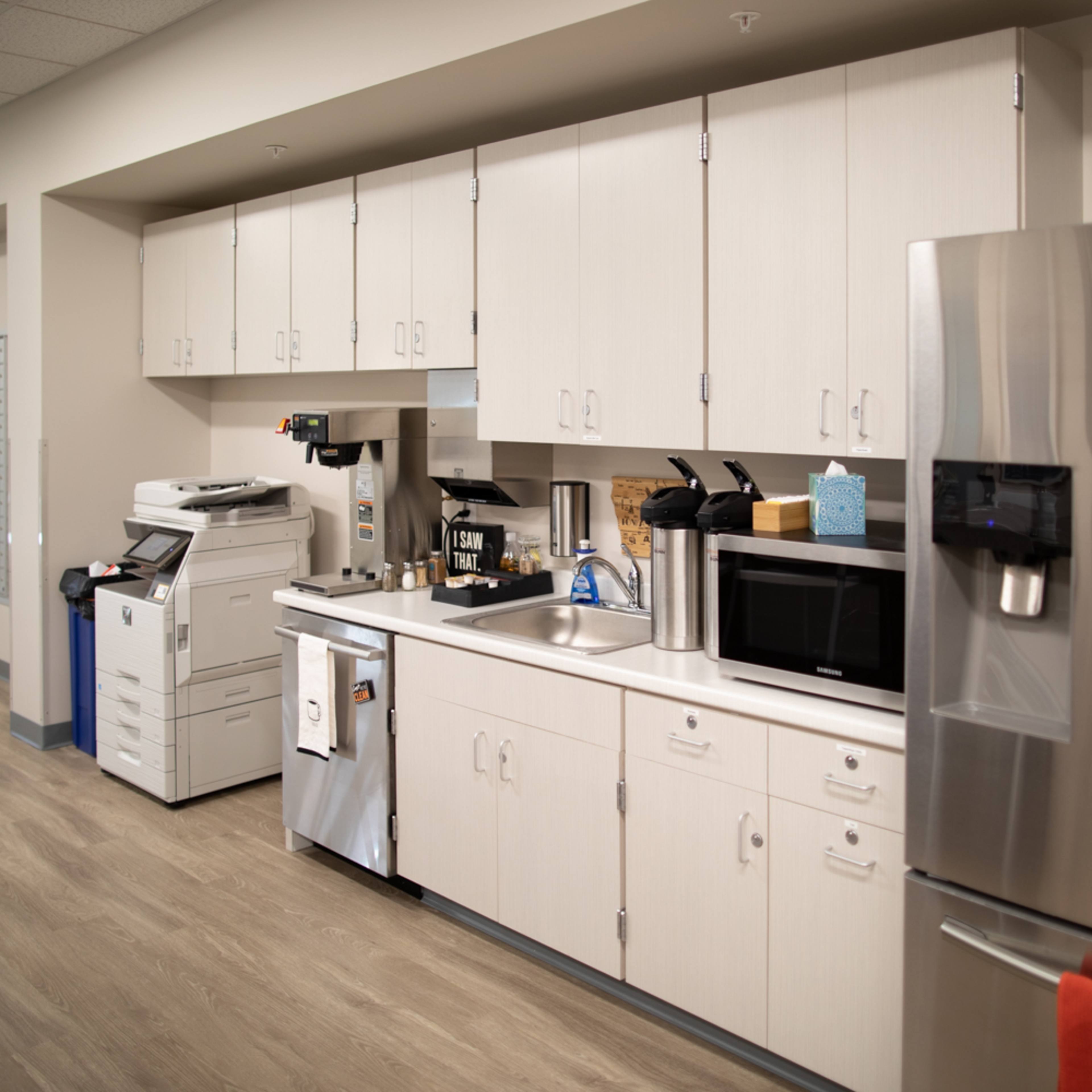 A modern office kitchen area with cabinets, a sink, a coffee machine, a microwave, and a stainless steel refrigerator.