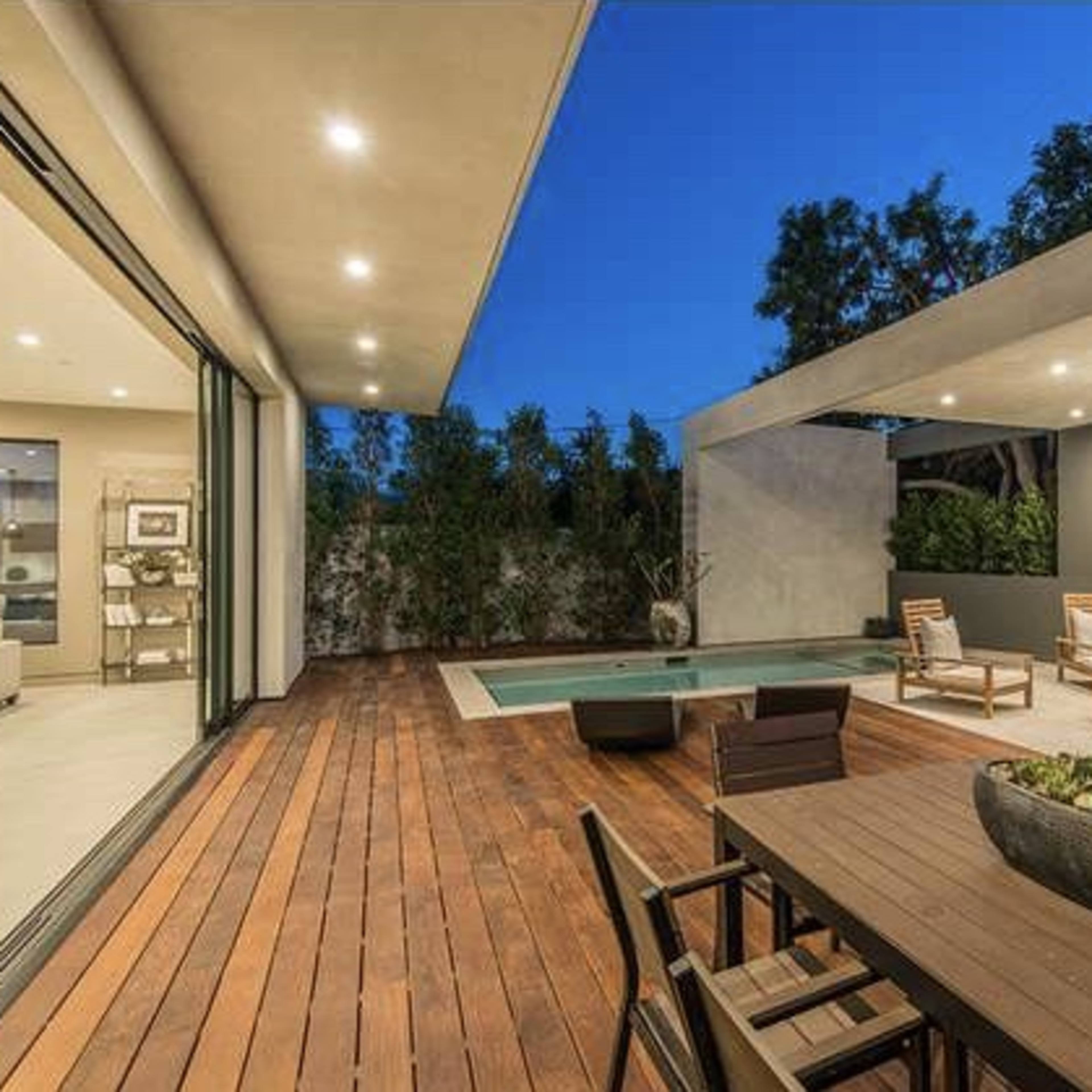 A modern outdoor patio with a wooden deck, a swimming pool, and sleek furniture under a softly lit overhead fixture.