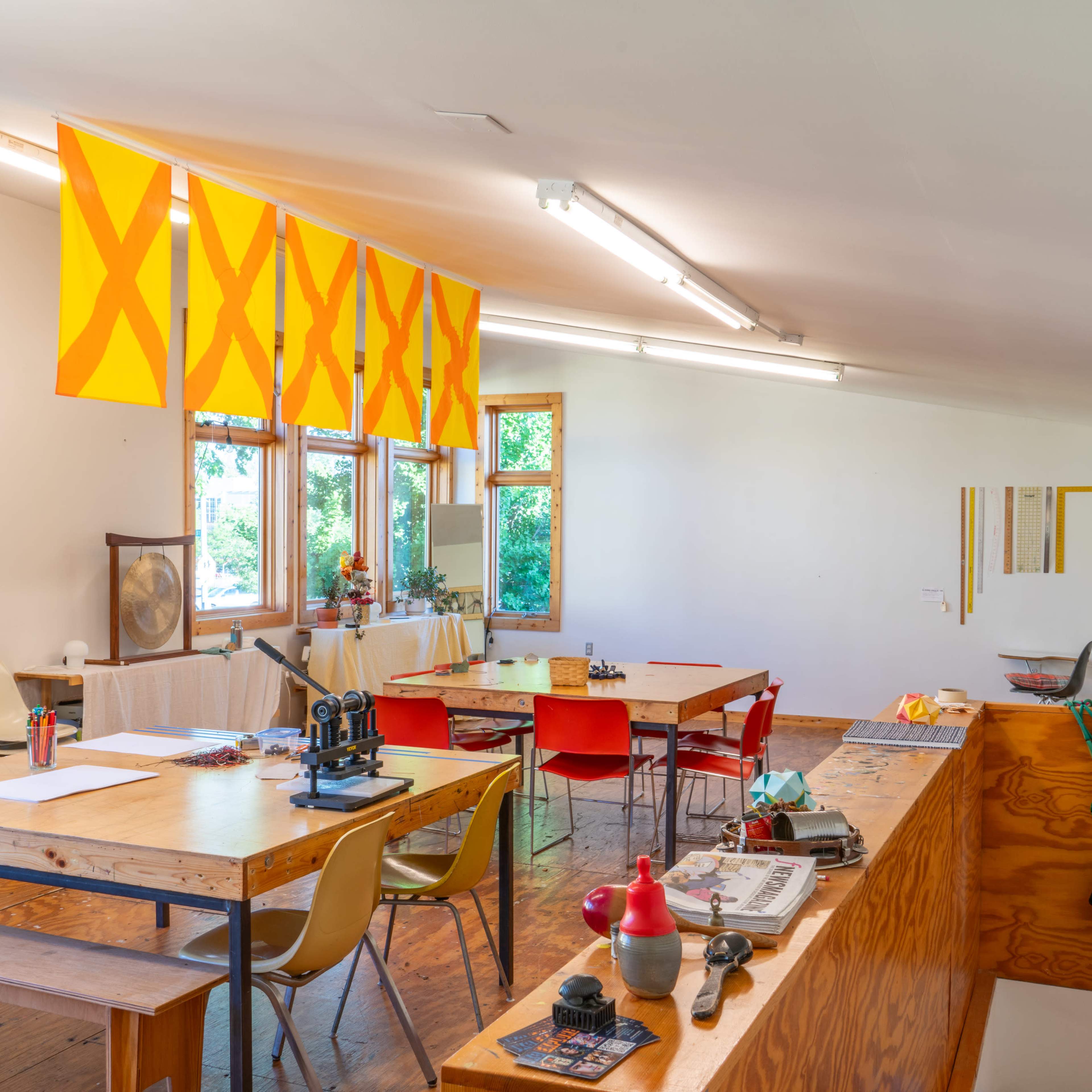 A bright, spacious room with several tables, colorful chairs, large windows, and yellow flags hanging from the ceiling.