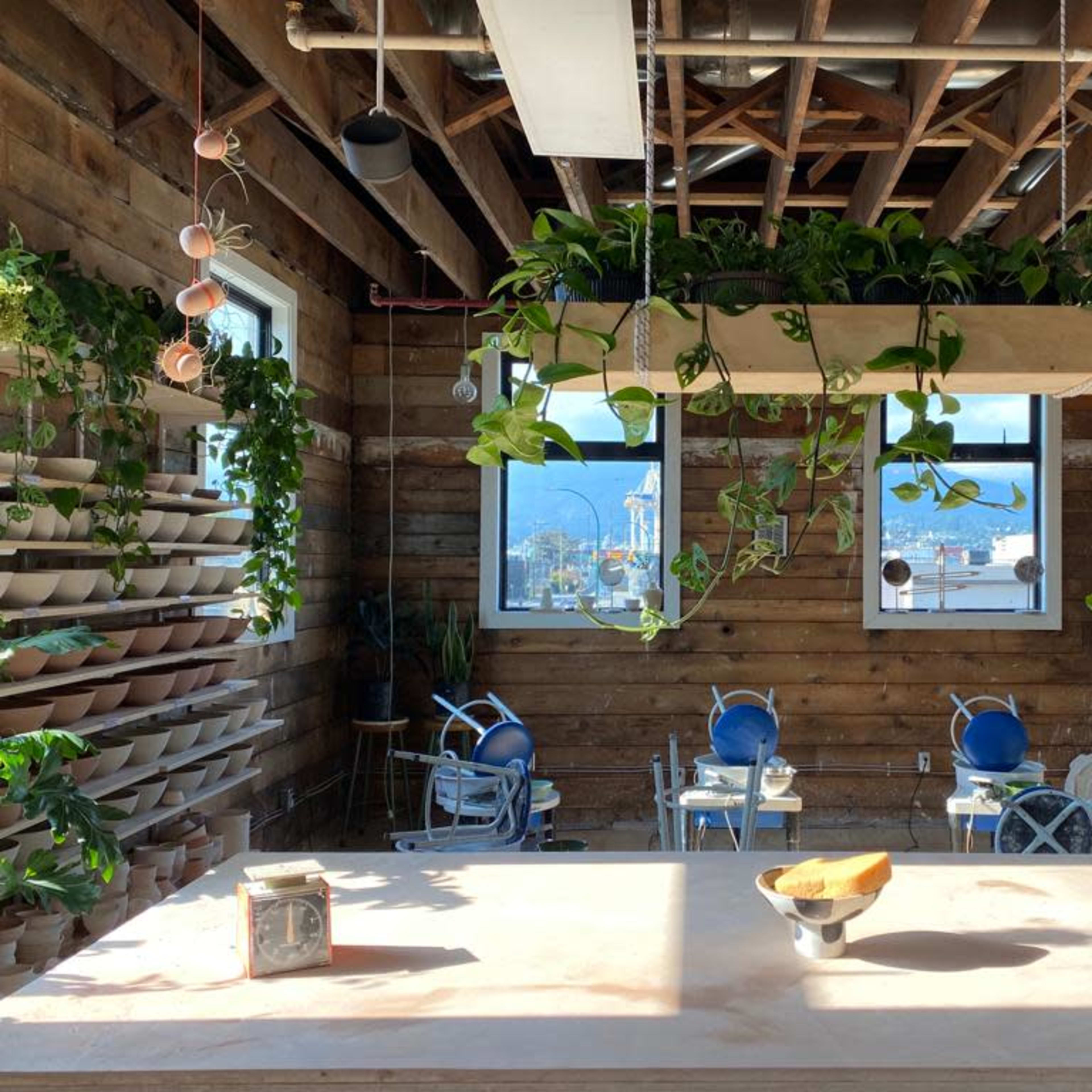 A spacious studio with wooden walls, hanging plants, and a large worktable in the foreground, overlooking a city view through large windows.