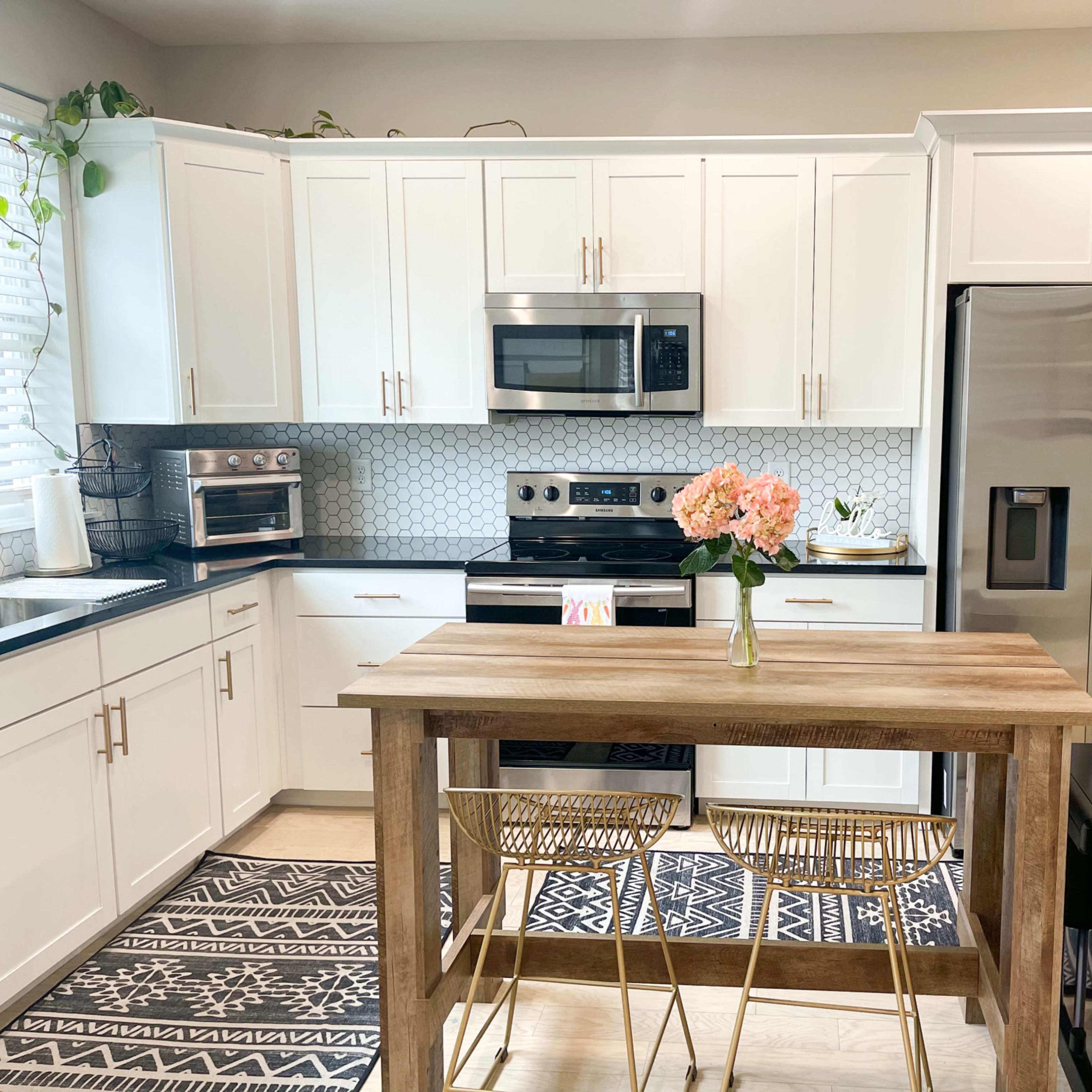 A modern kitchen features white cabinets, stainless steel appliances, and a wooden table with two chairs, complemented by a decorative plant and flowers.