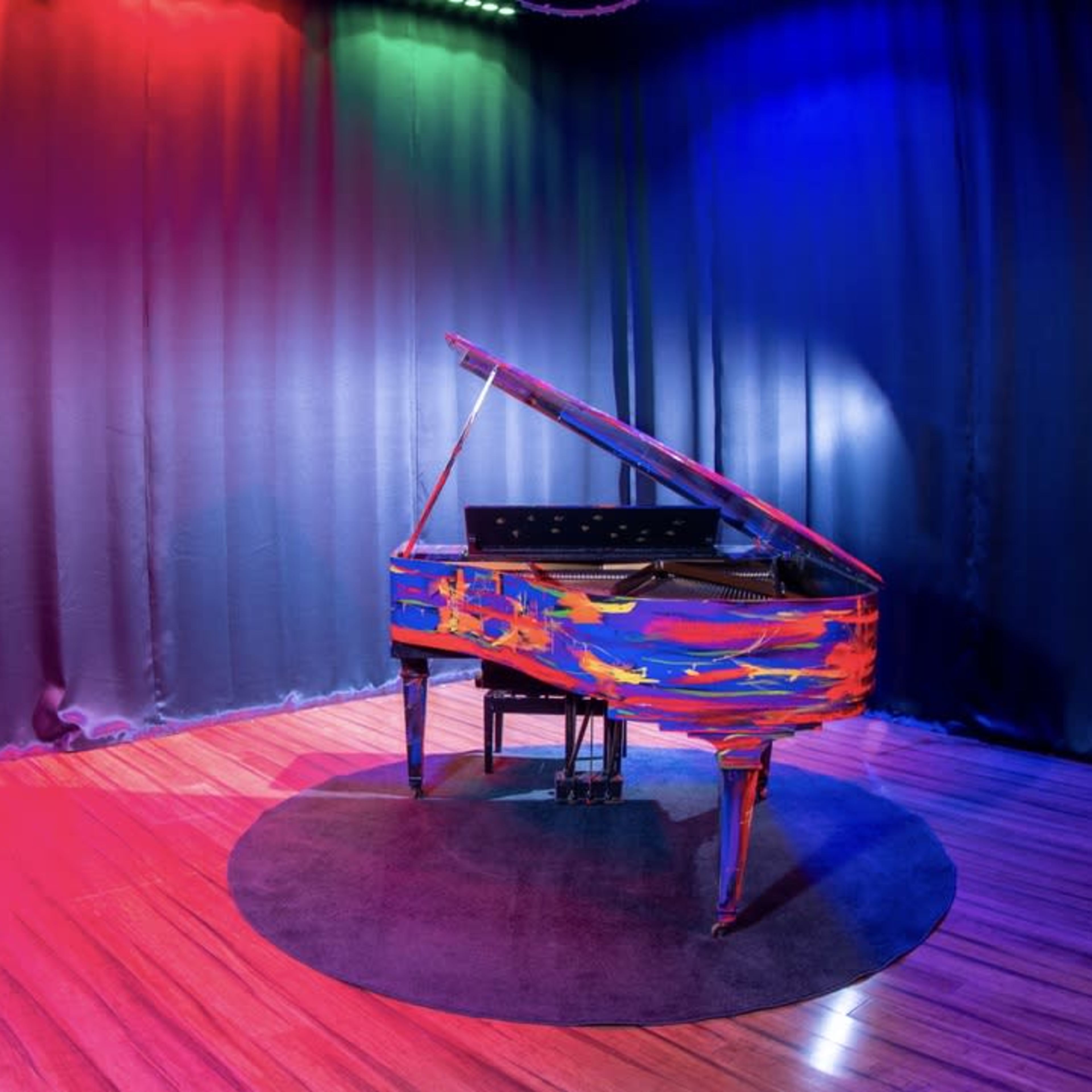 A brightly painted grand piano sits on a circular rug in a stage area illuminated by colorful lights against a dark backdrop.