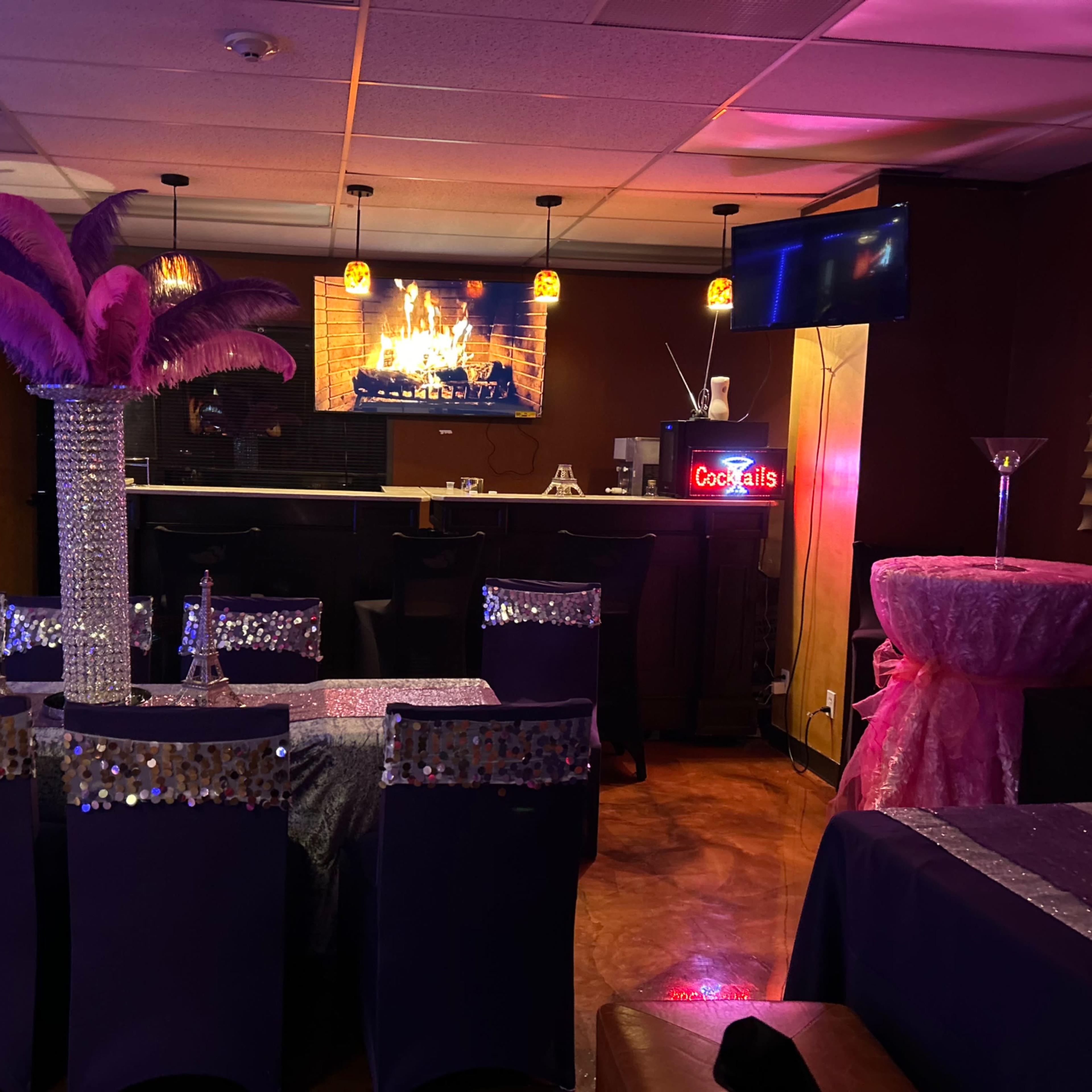 A dimly lit event space with purple decor, a bar in the background, and a cozy ambiance created by a faux fireplace on the screen.