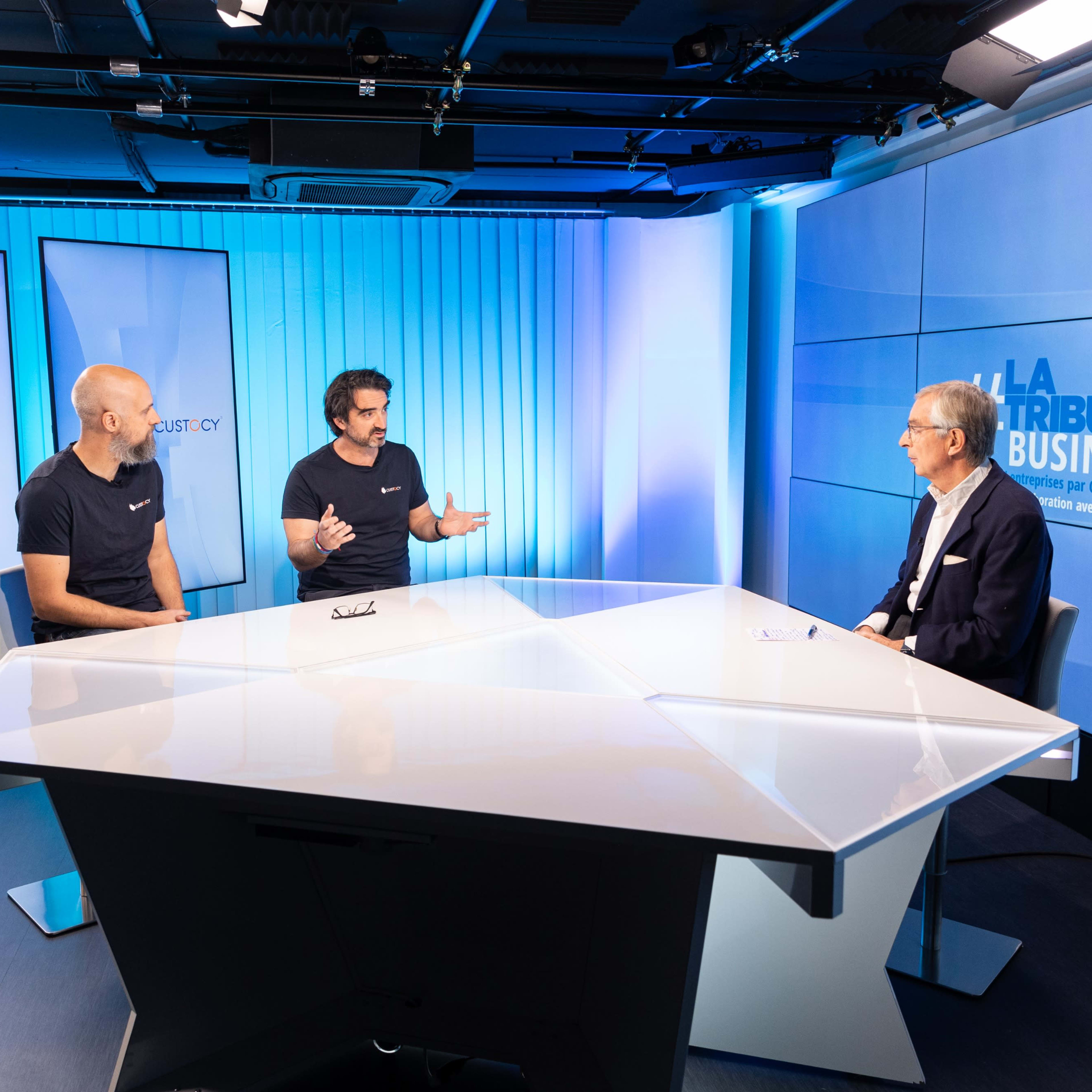 Three men are seated at a white table in a studio, discussing in front of screens displaying "LA TRIBUNE BUSINESS."