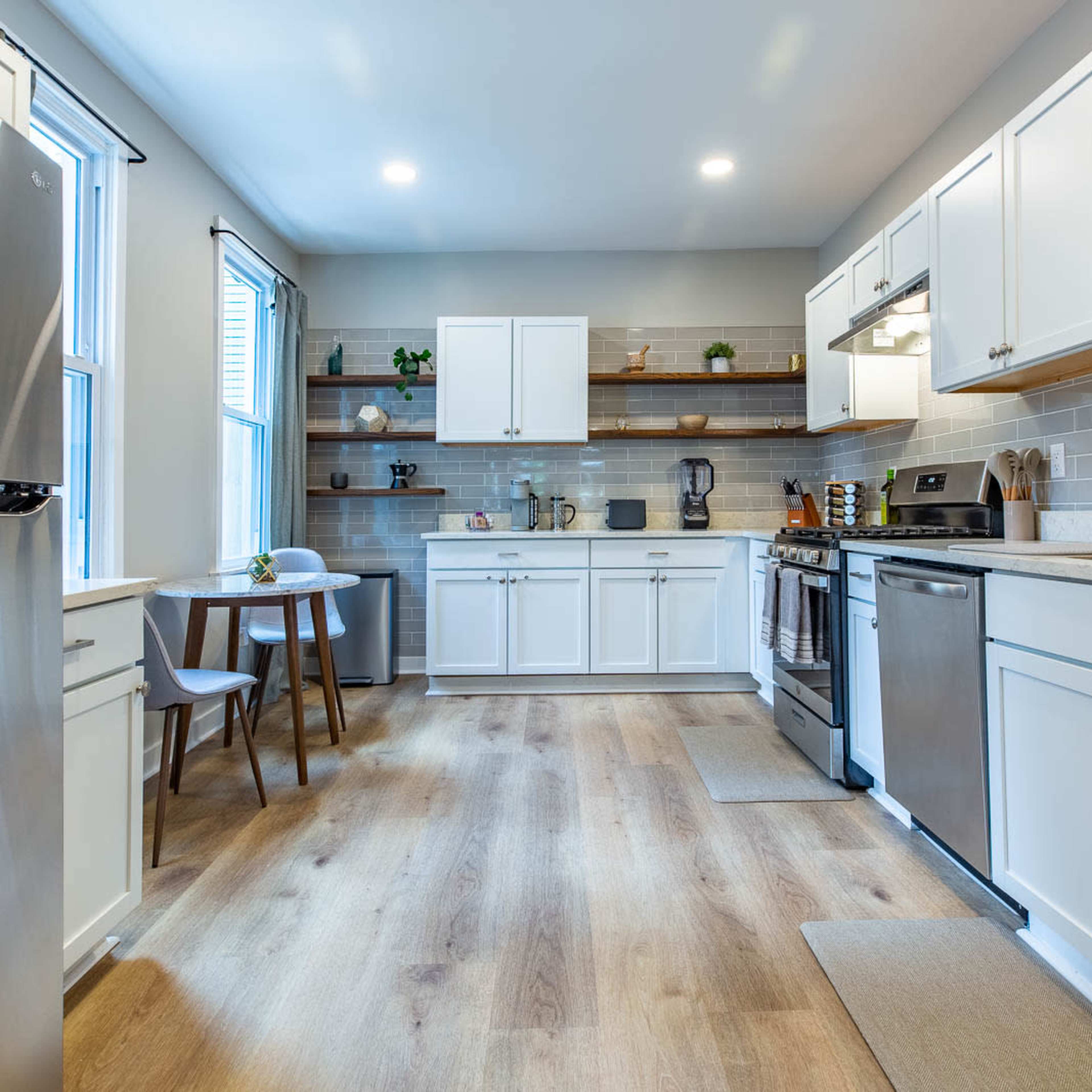 A modern kitchen featuring stainless steel appliances, white cabinetry, and wooden flooring with a small dining table and two chairs beside a window.