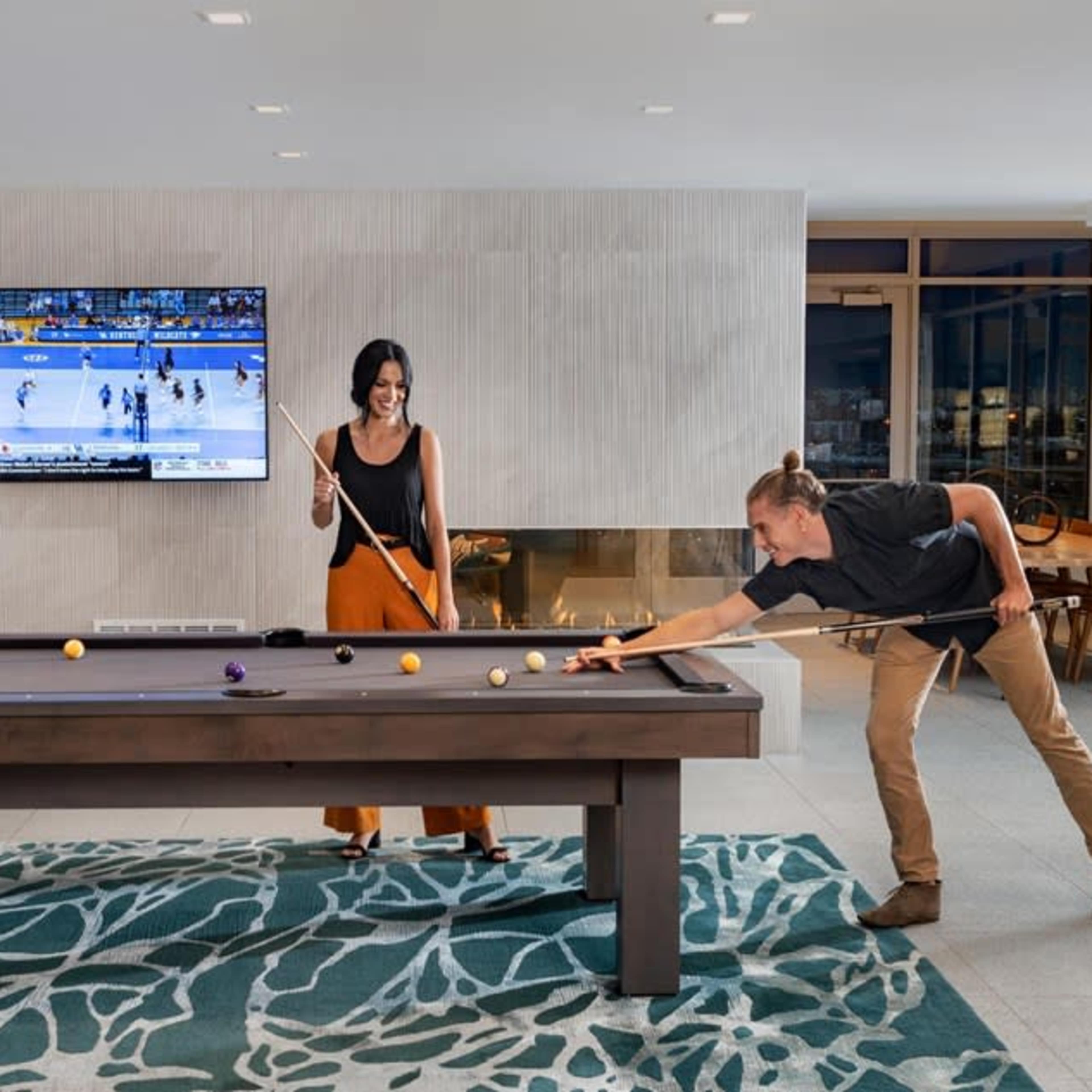 A man and a woman play billiards in a modern room with large windows and a television displaying a sports game.