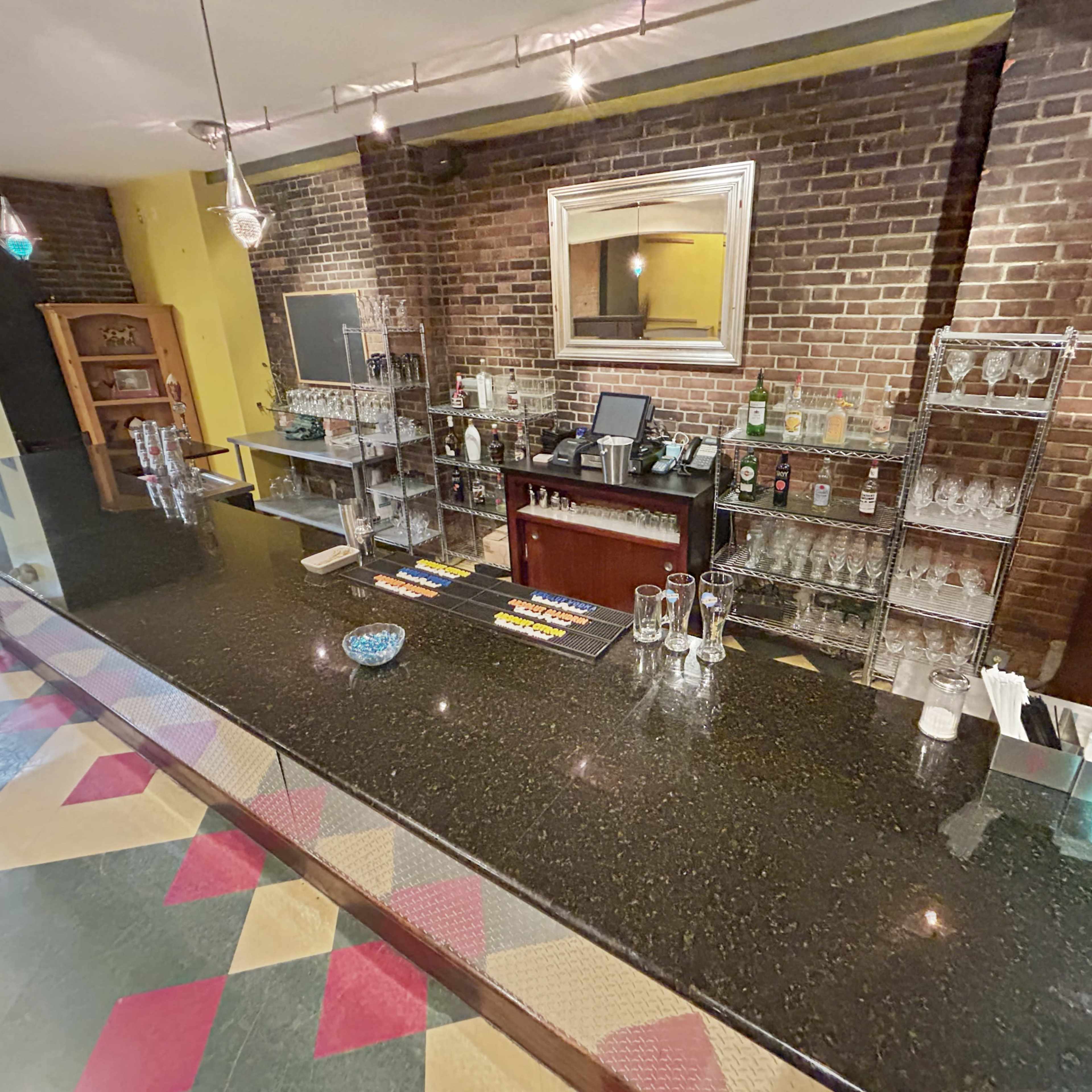 A bar area with a dark countertop, shelves displaying various bottles and glassware, and a brick wall in the background.