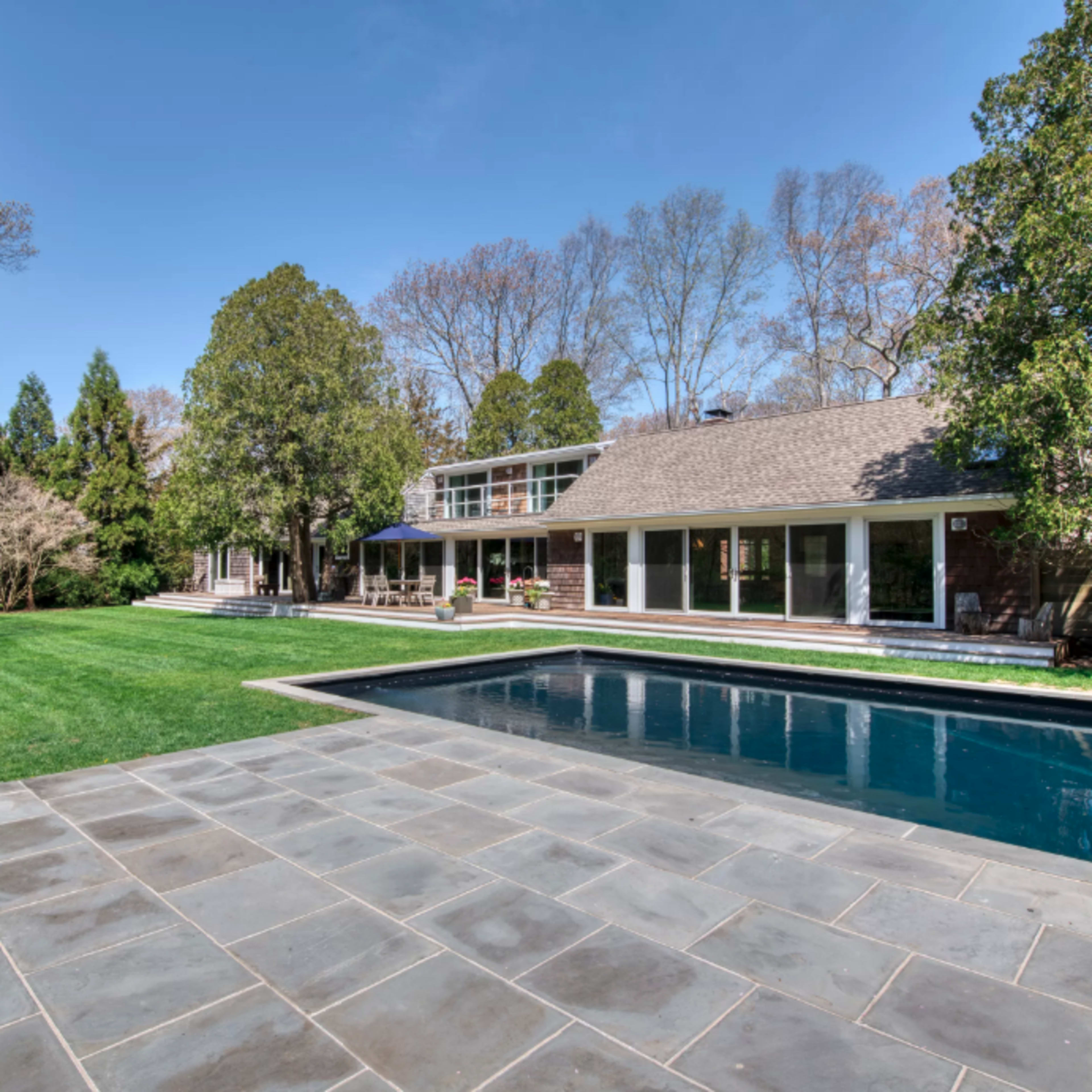 A modern house with a stone patio and a swimming pool, surrounded by a well-kept lawn and trees in the background.