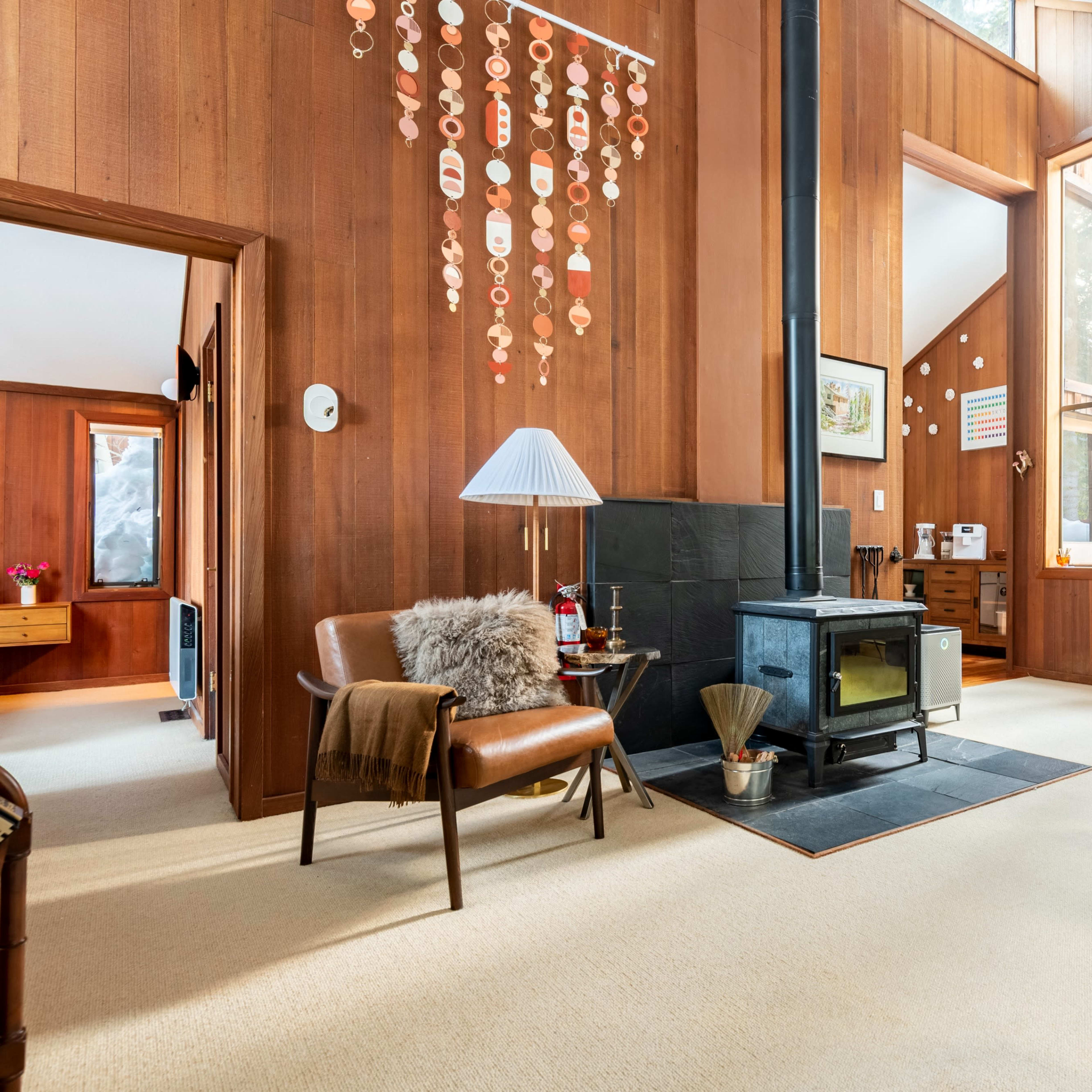 A wooden-paneled living room with a wood stove, a cozy armchair, and large windows allowing natural light to enter.