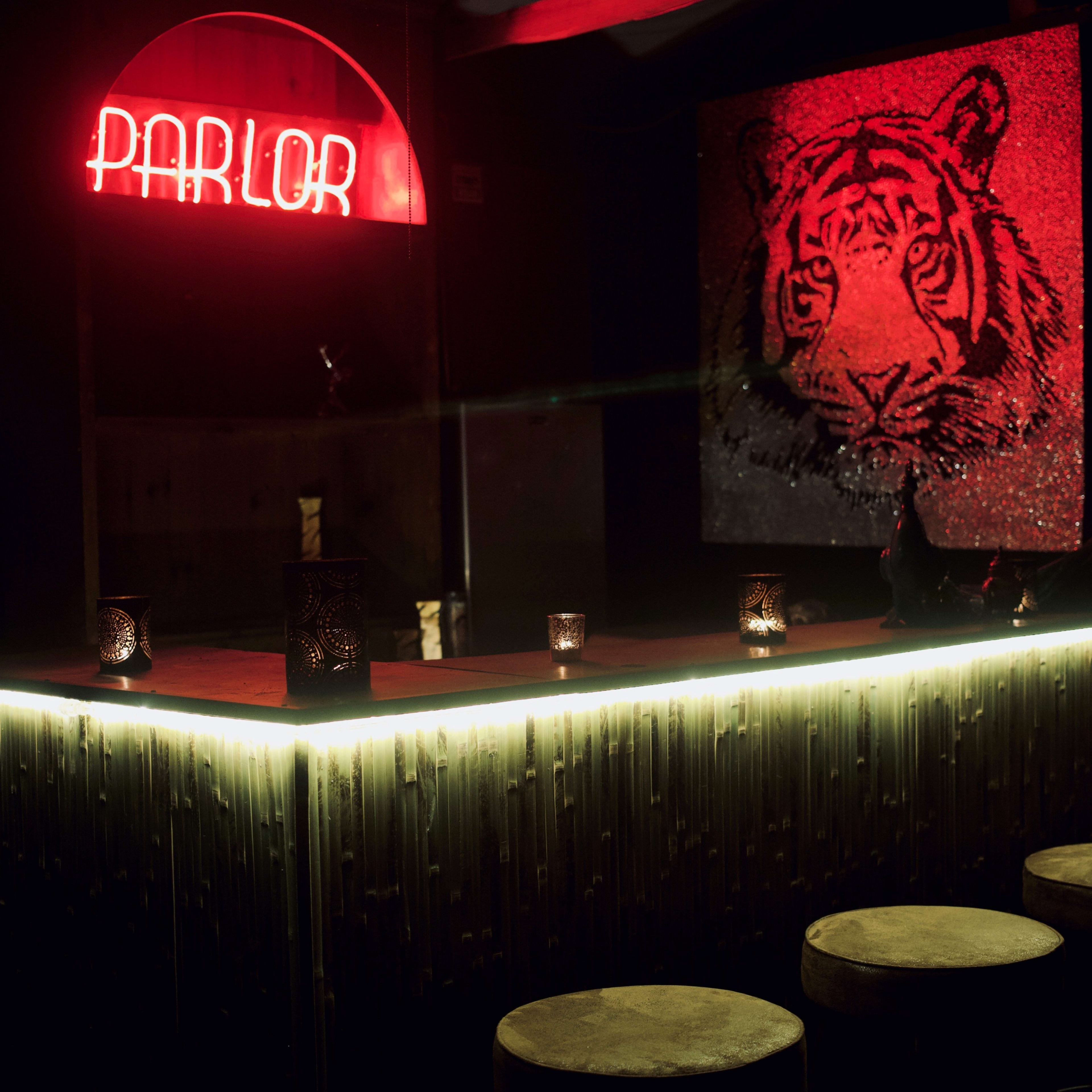 A dimly lit bar with a neon "PARLOR" sign and a large tiger portrait on the wall features wooden stools and candles on the counter.