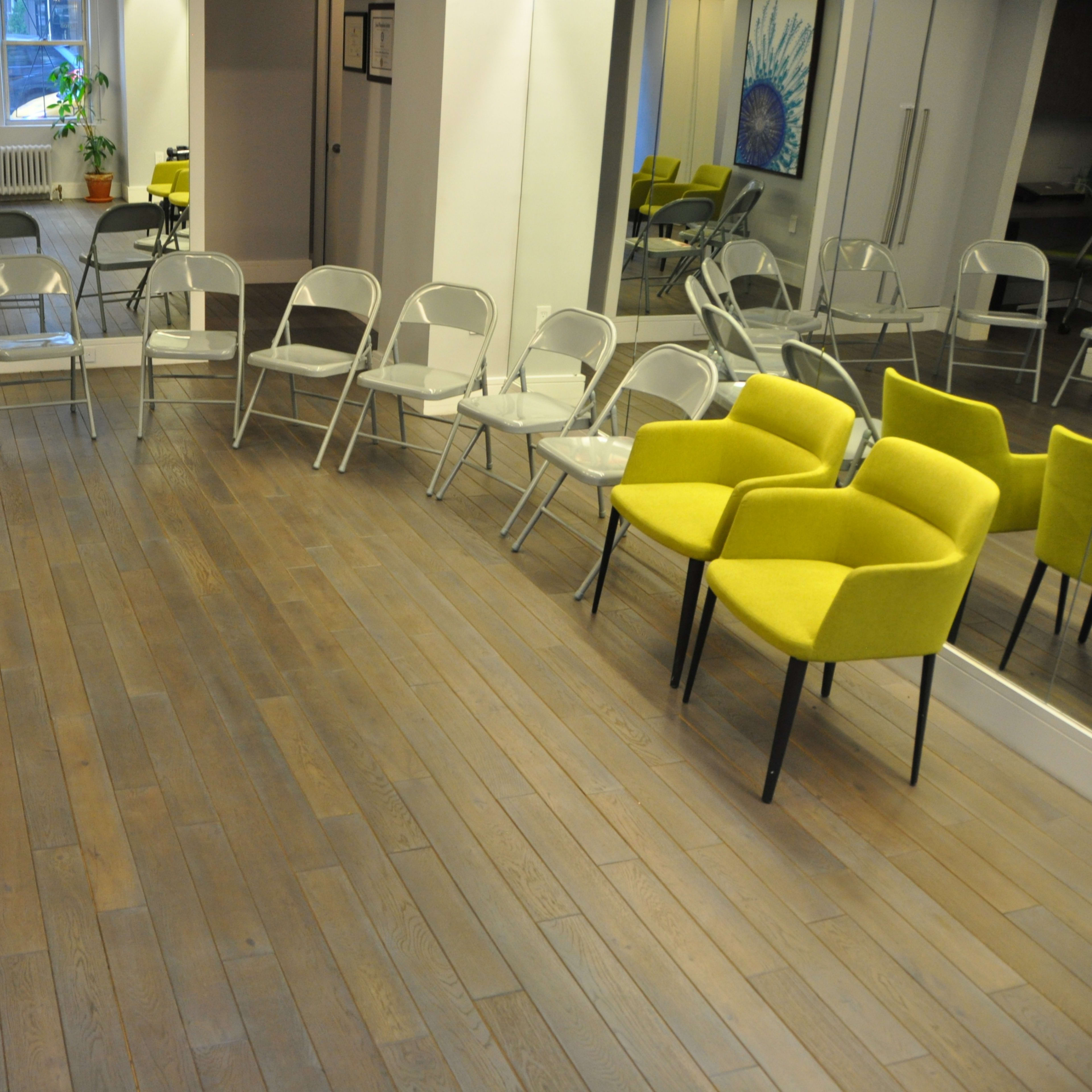 A spacious room with wooden flooring, featuring yellow armchairs and transparent folding chairs arranged in a circular setup around mirrored walls.