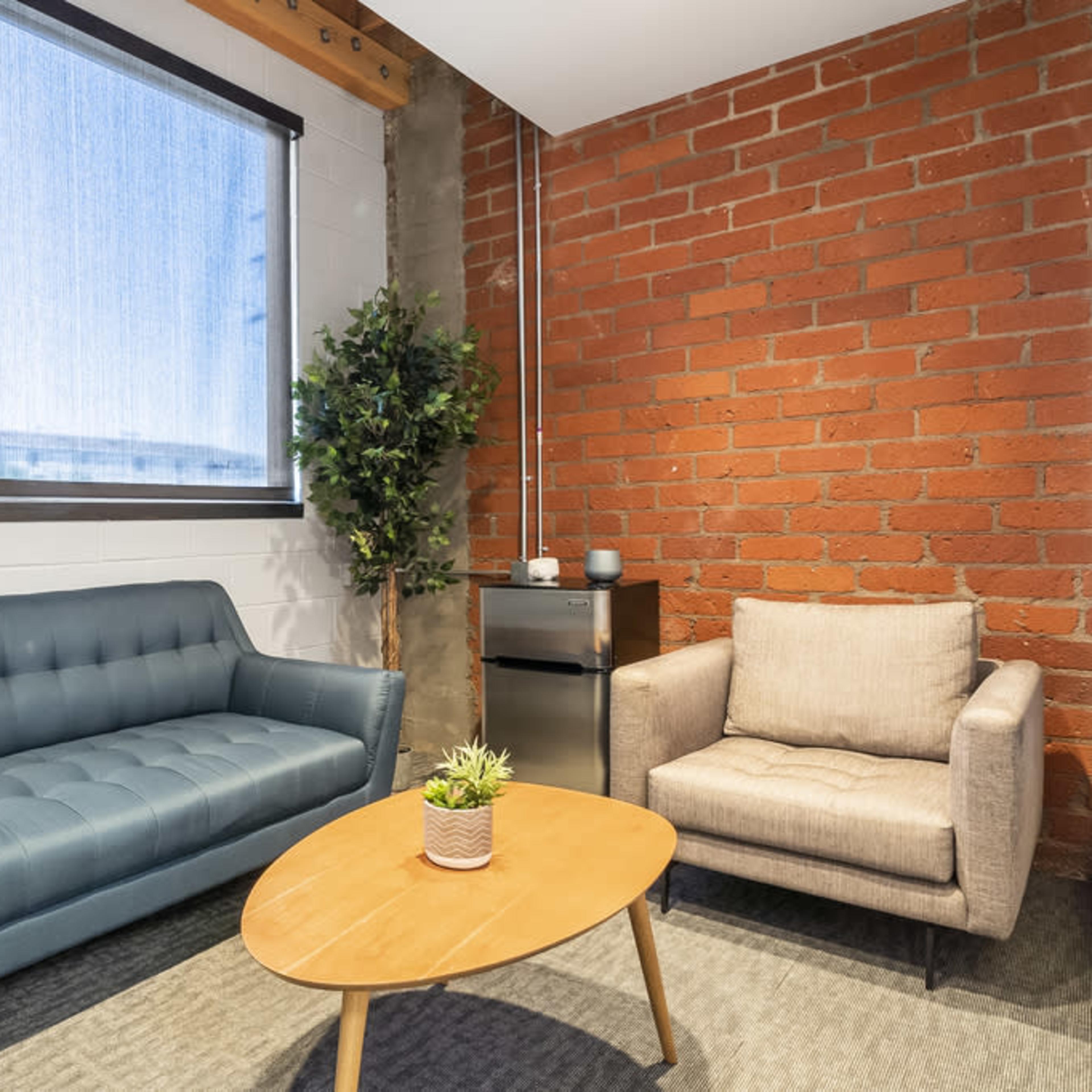 A small modern lounge area featuring a blue sofa, a beige armchair, a wooden coffee table, and a potted plant, with a large window allowing natural light to enter.
