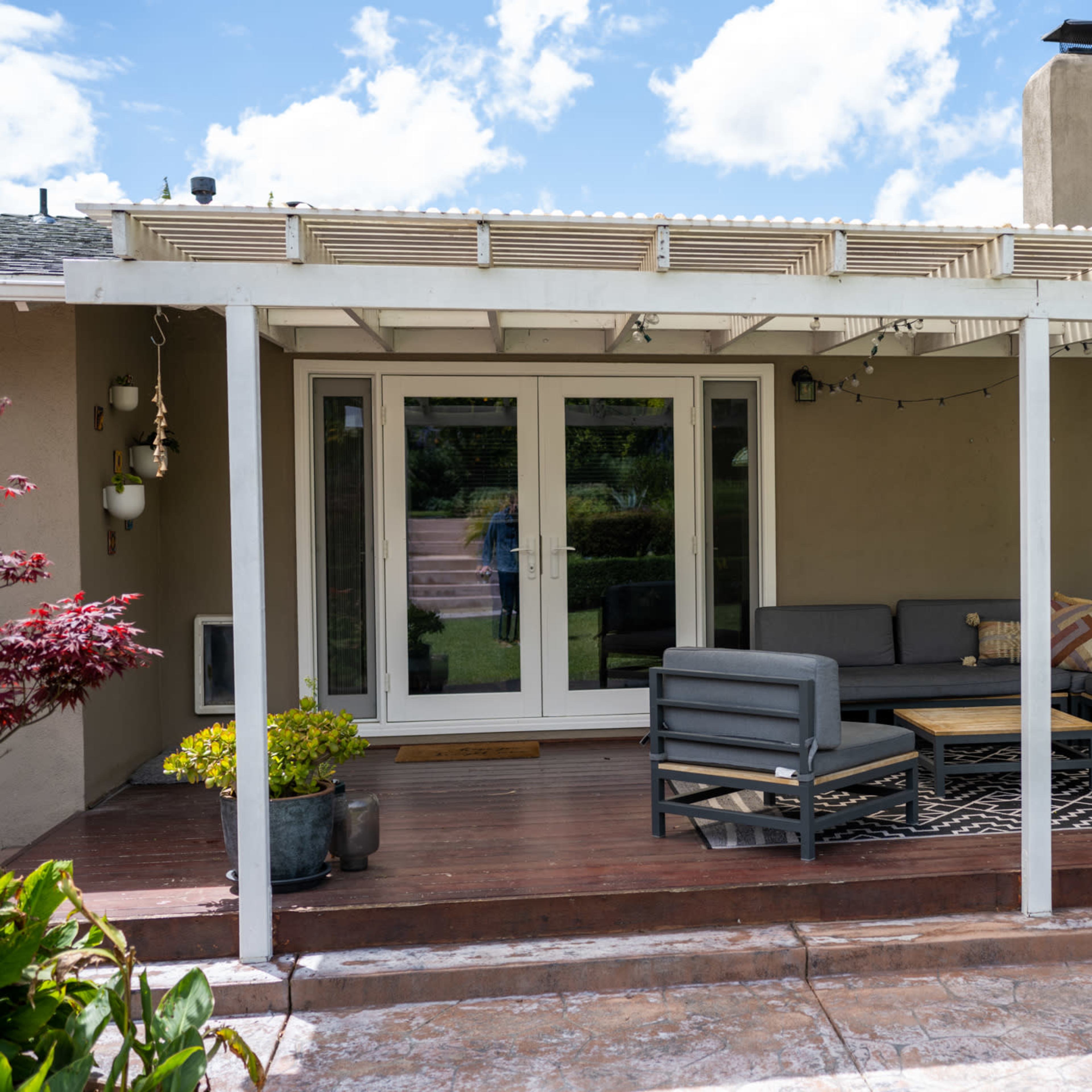 A covered patio features a seating area with a sectional sofa and a coffee table, surrounded by potted plants and a decorative wind chime.