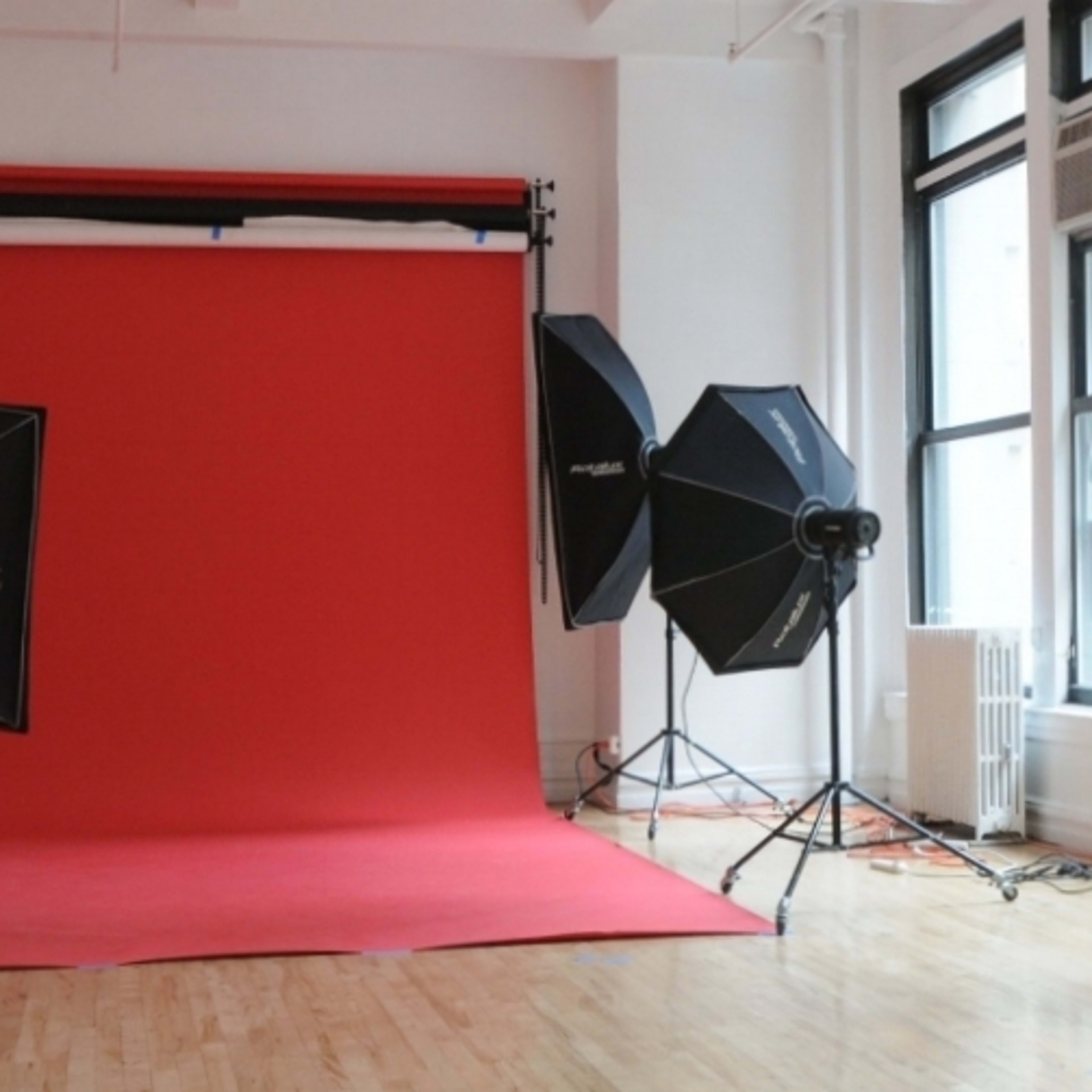 A photography studio with a red backdrop, various lighting equipment, and large windows allowing natural light to enter.