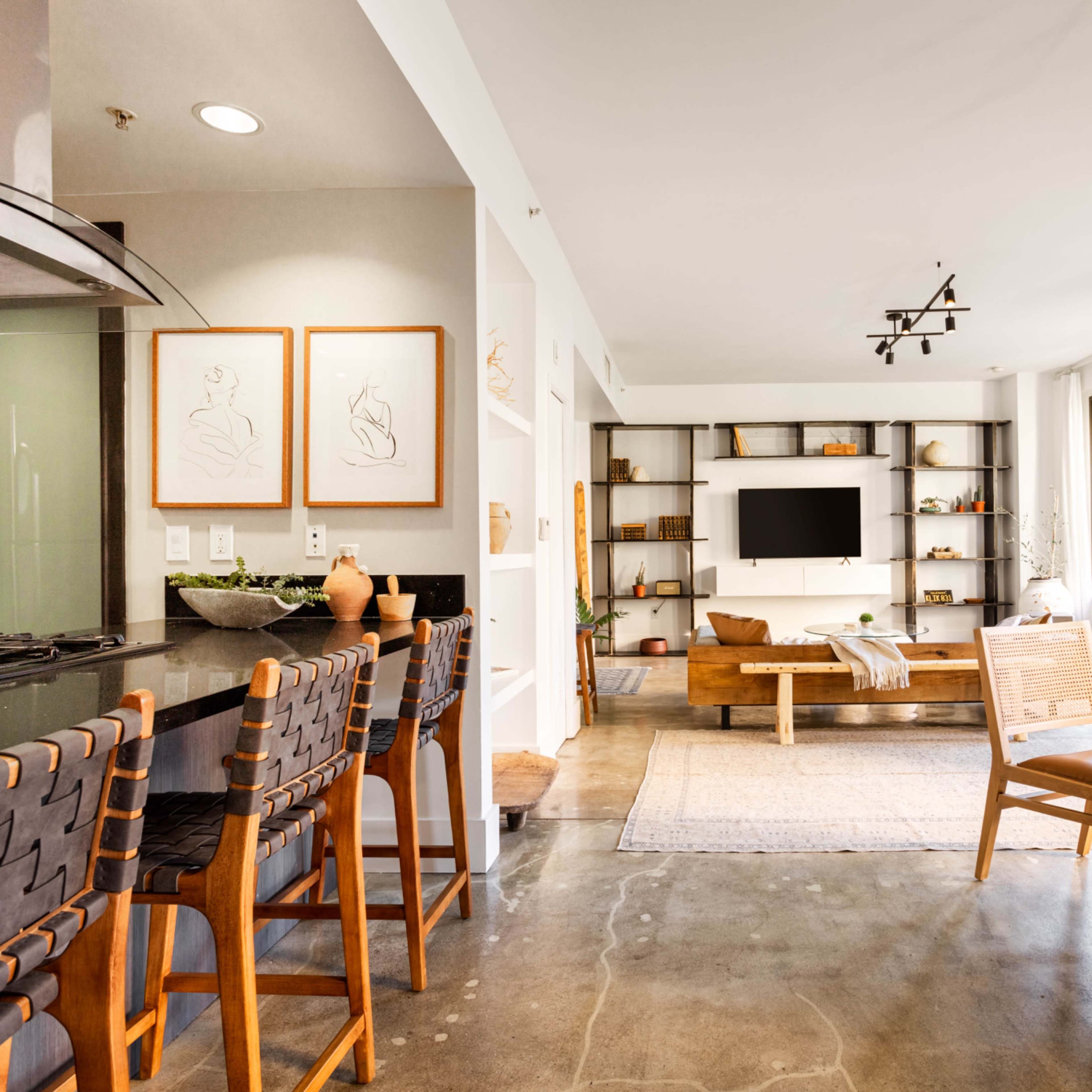 A modern living space with a kitchen bar, a dining table, and a seating area featuring a television and shelves against a wall of large windows.