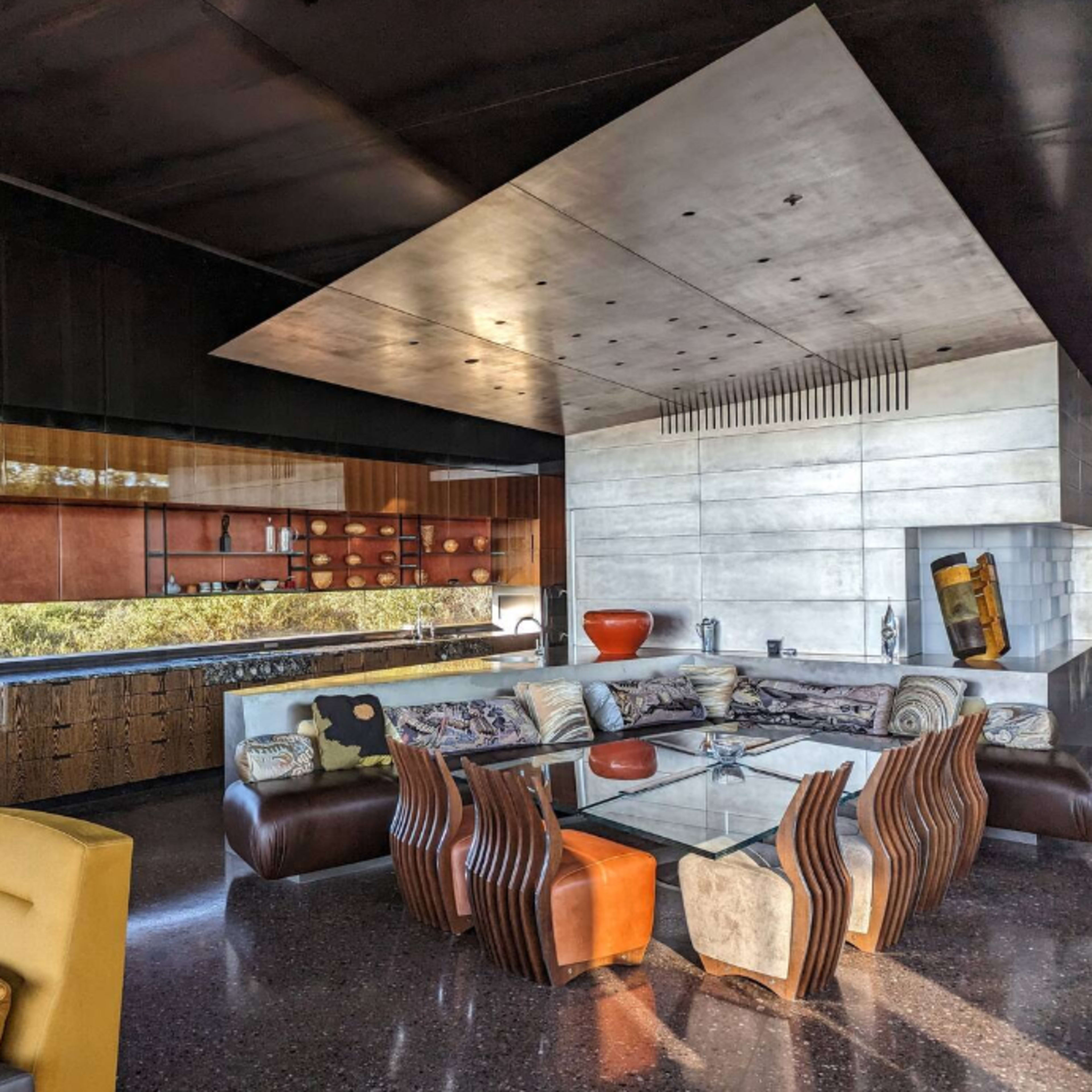 A modern living room with a glass table surrounded by stylish seating, featuring a sleek kitchen area with a contrast of materials and a view of greenery through large windows.