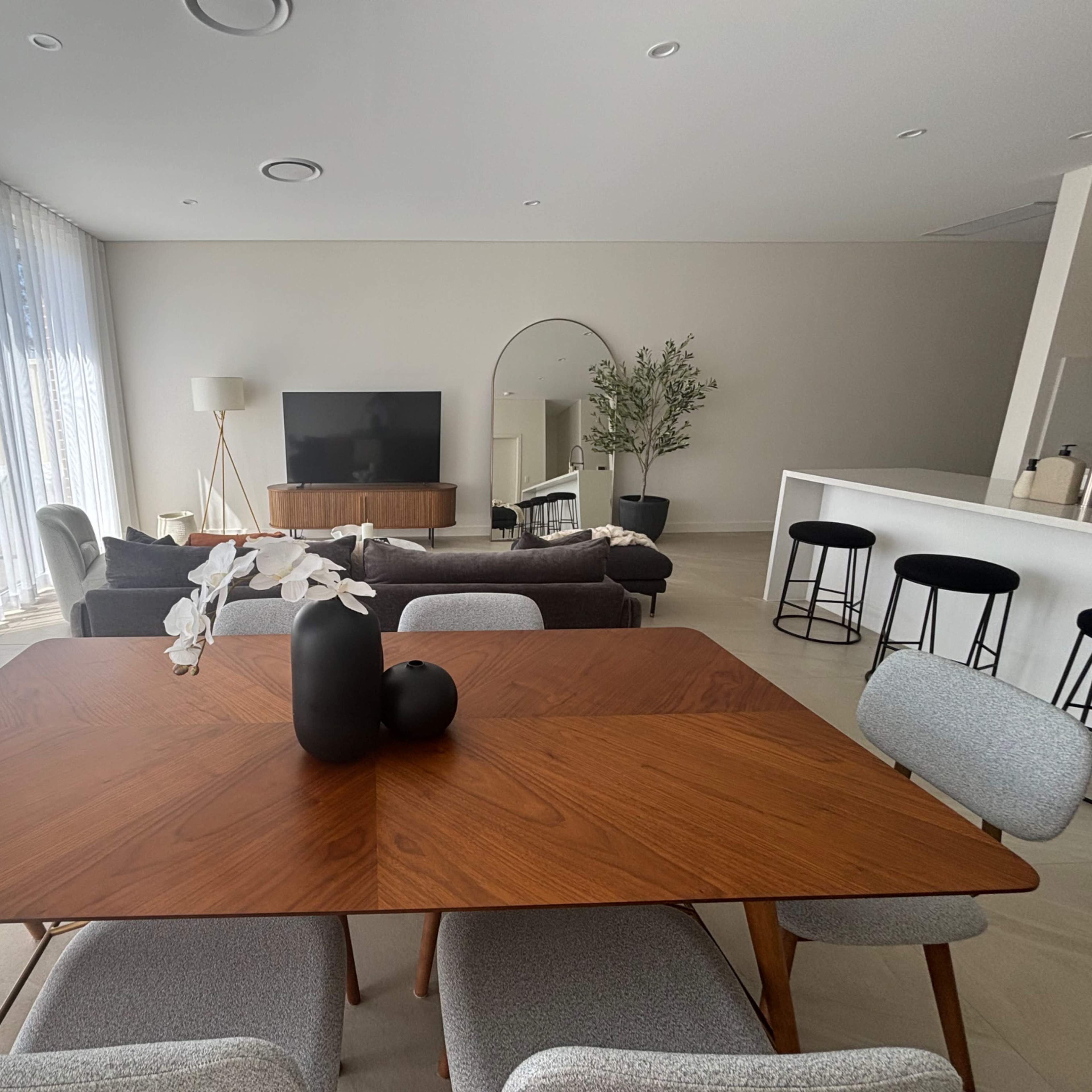 A modern living space features a wooden dining table with vases at its center, surrounded by upholstered chairs, a sofa area, and a television mounted on the wall.
