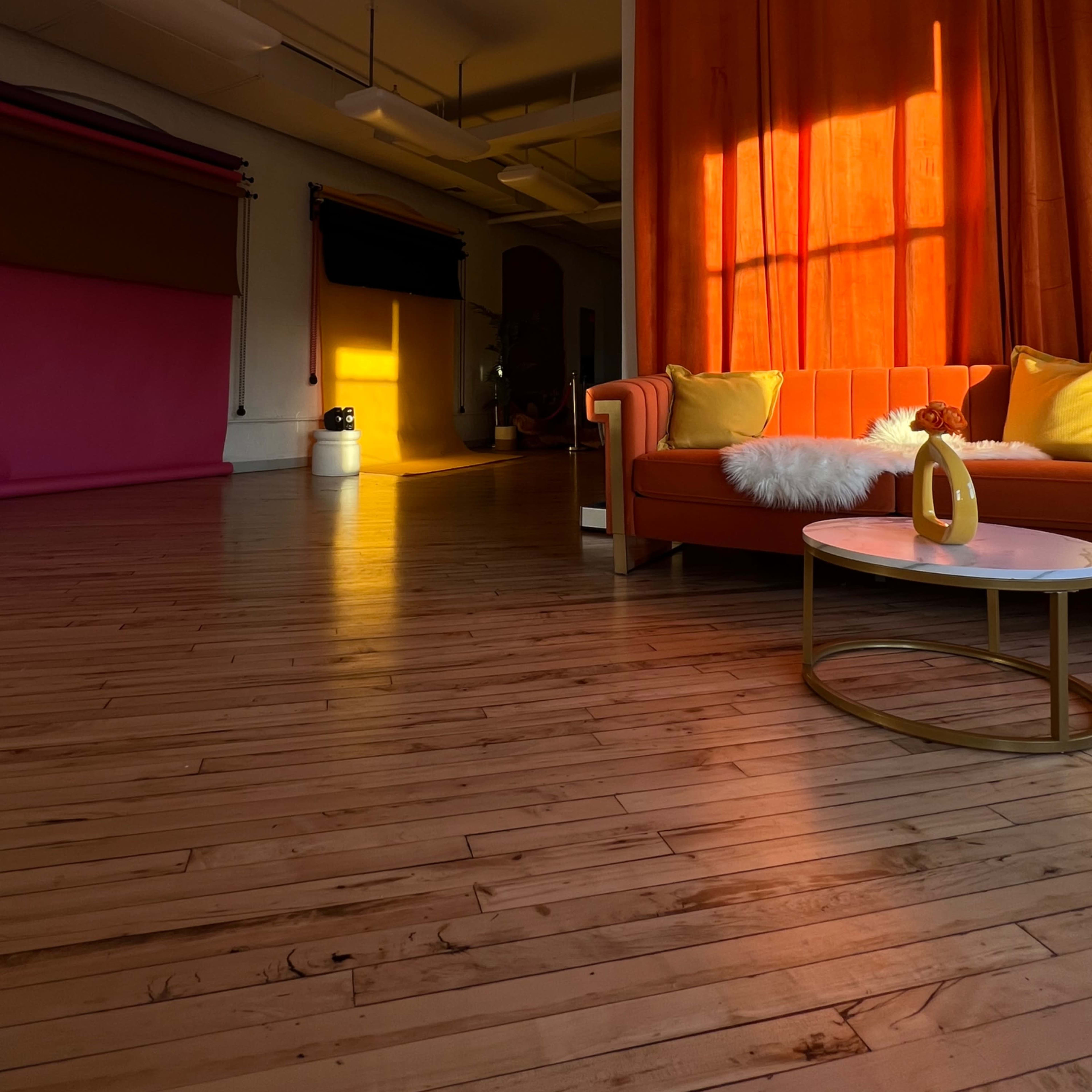 A brightly lit room features an orange curtain, a pink backdrop, a wooden floor, and a cozy orange sofa with a white pillow and a round table.