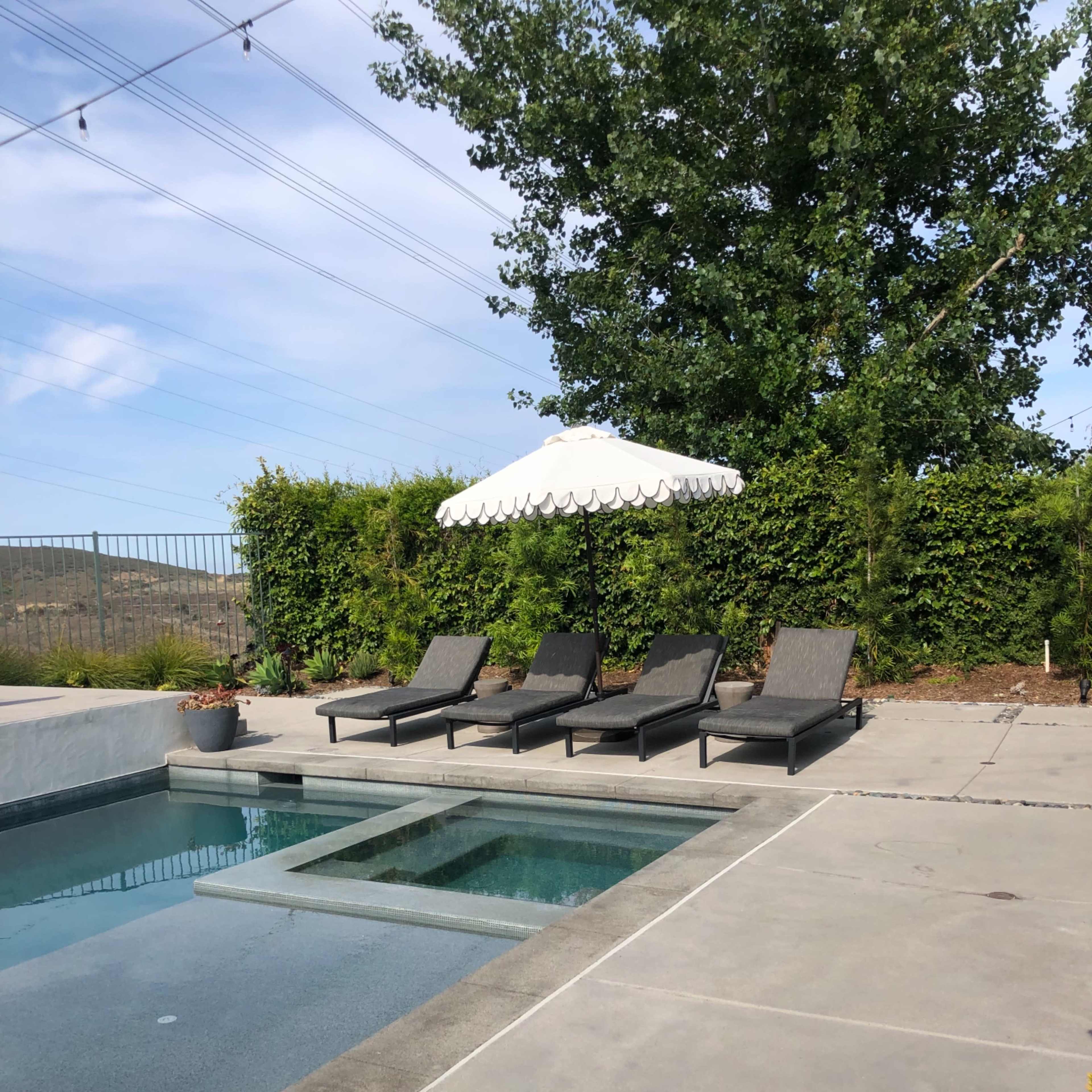 A pool area features four lounge chairs under a large umbrella beside a clear water pool.