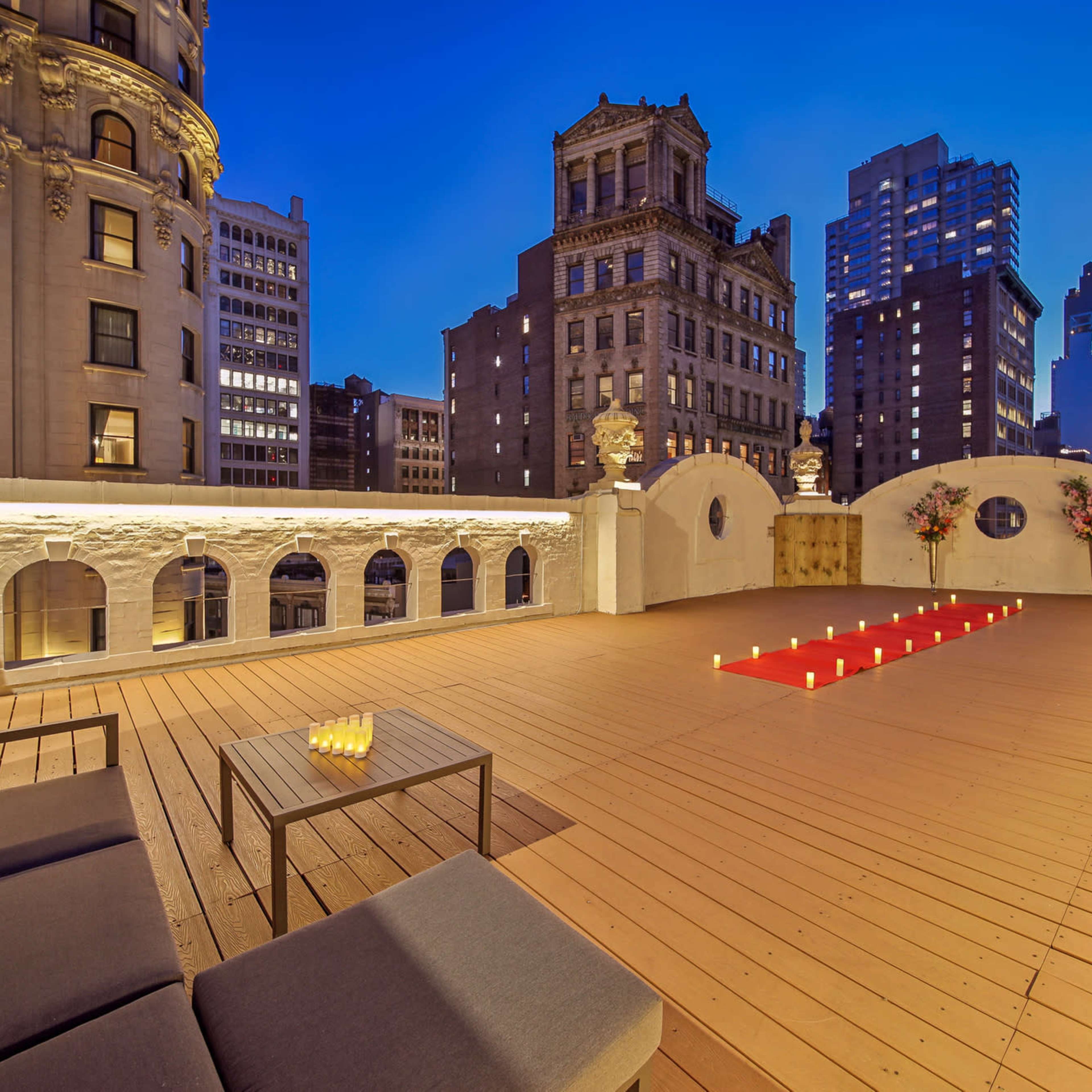 A spacious rooftop terrace with wooden flooring, featuring modern seating, lit candles, and a view of city buildings at dusk.