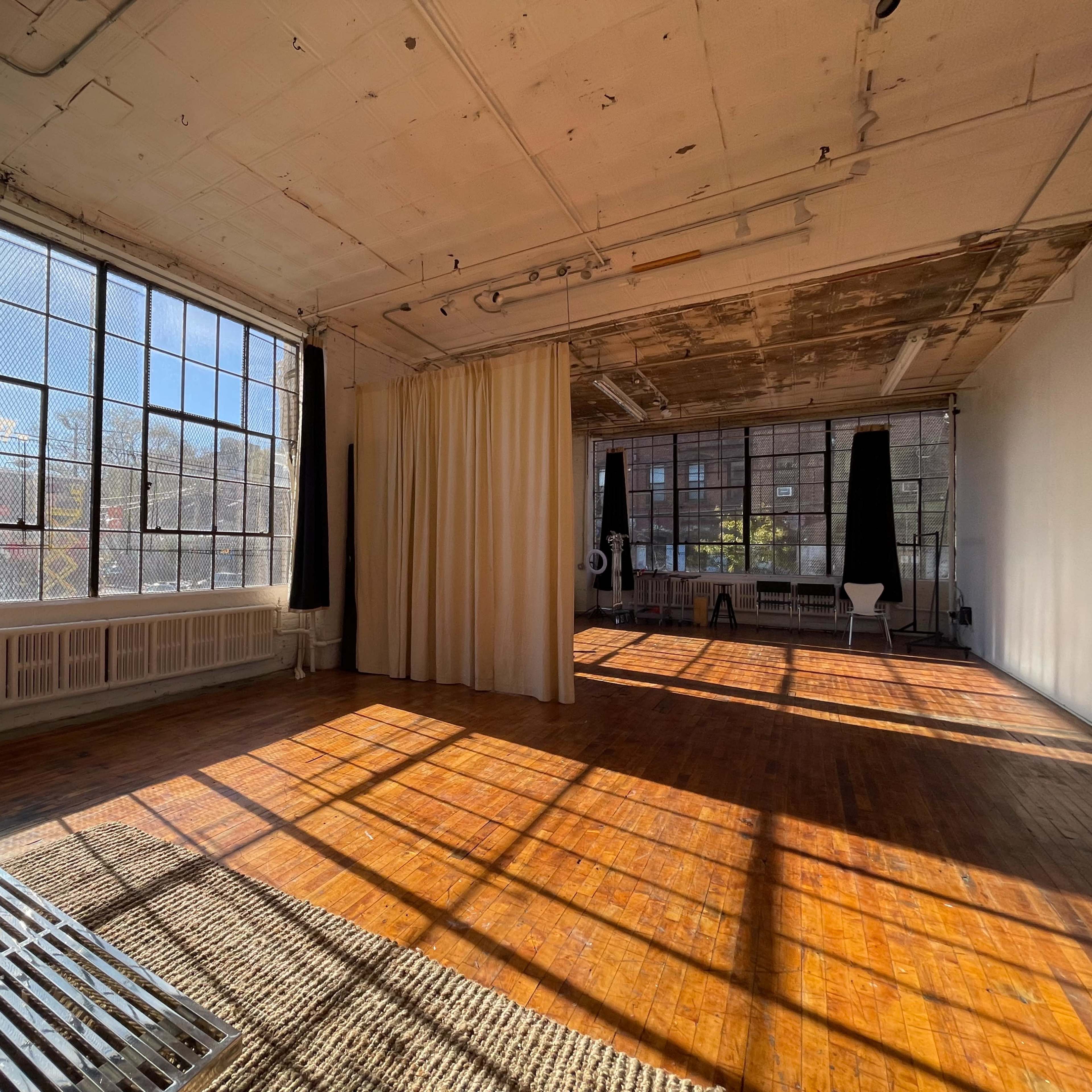 A sunlit, spacious room with large windows, wooden floors, and light-colored walls, featuring a seating area with a bench and curtains.