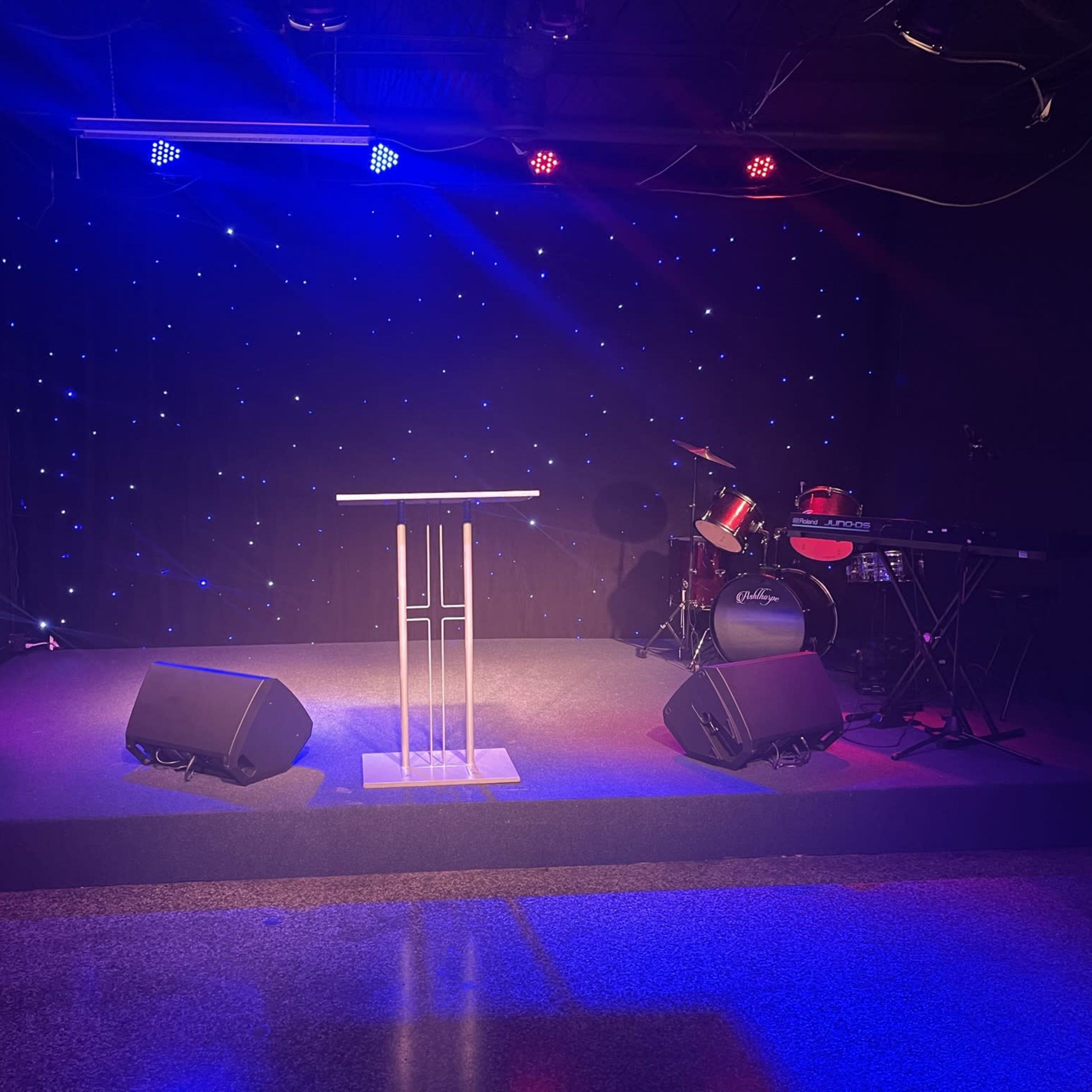 A performance stage featuring a microphone stand, a drum set, a keyboard, and illuminated by colored lights against a black backdrop with twinkling stars.