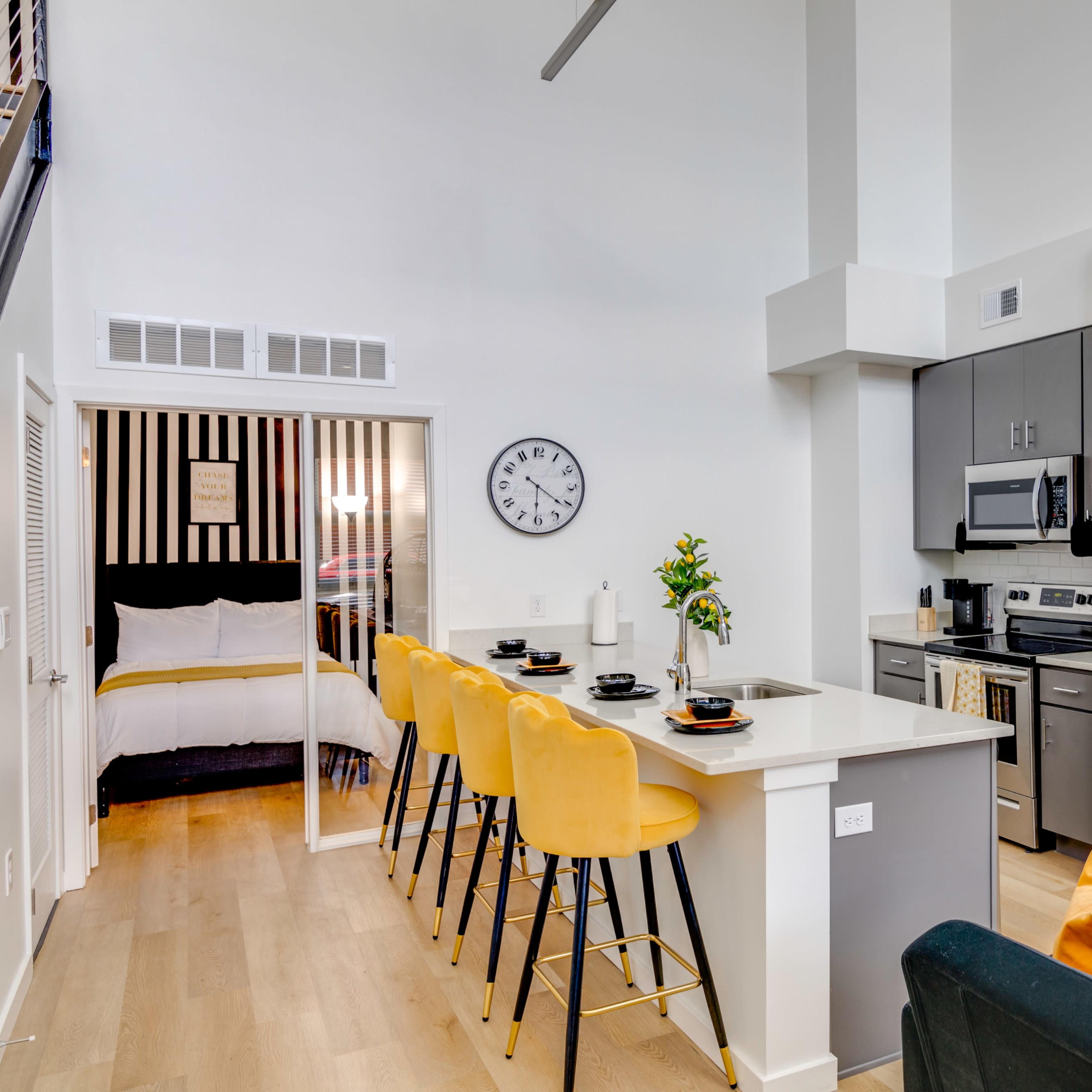 A modern kitchen area with stainless steel appliances, a breakfast bar with yellow stools, and a view into a bedroom with a black and white striped feature wall.