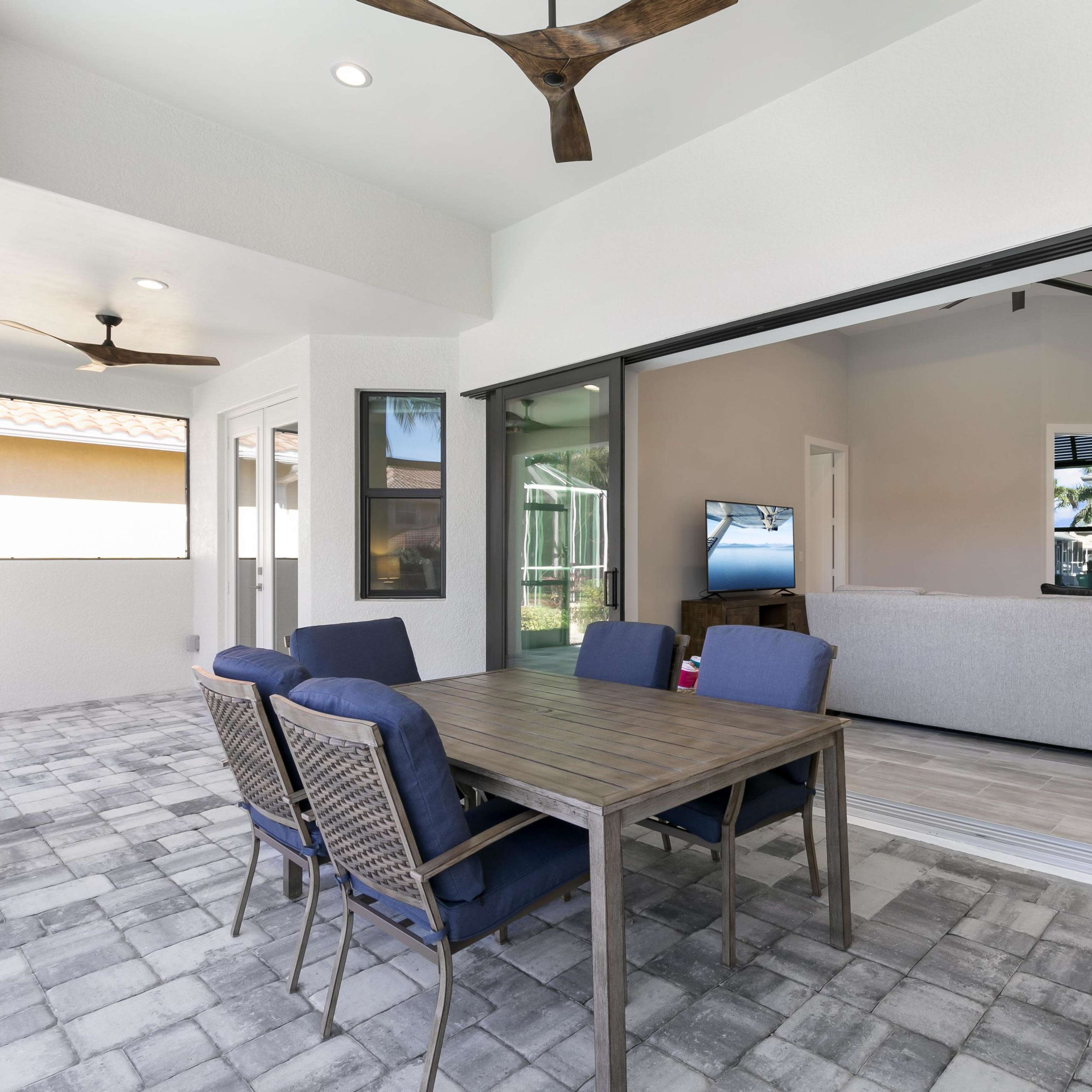 A patio area features a wooden dining table with four chairs, overlooking an indoor living space through large sliding glass doors.