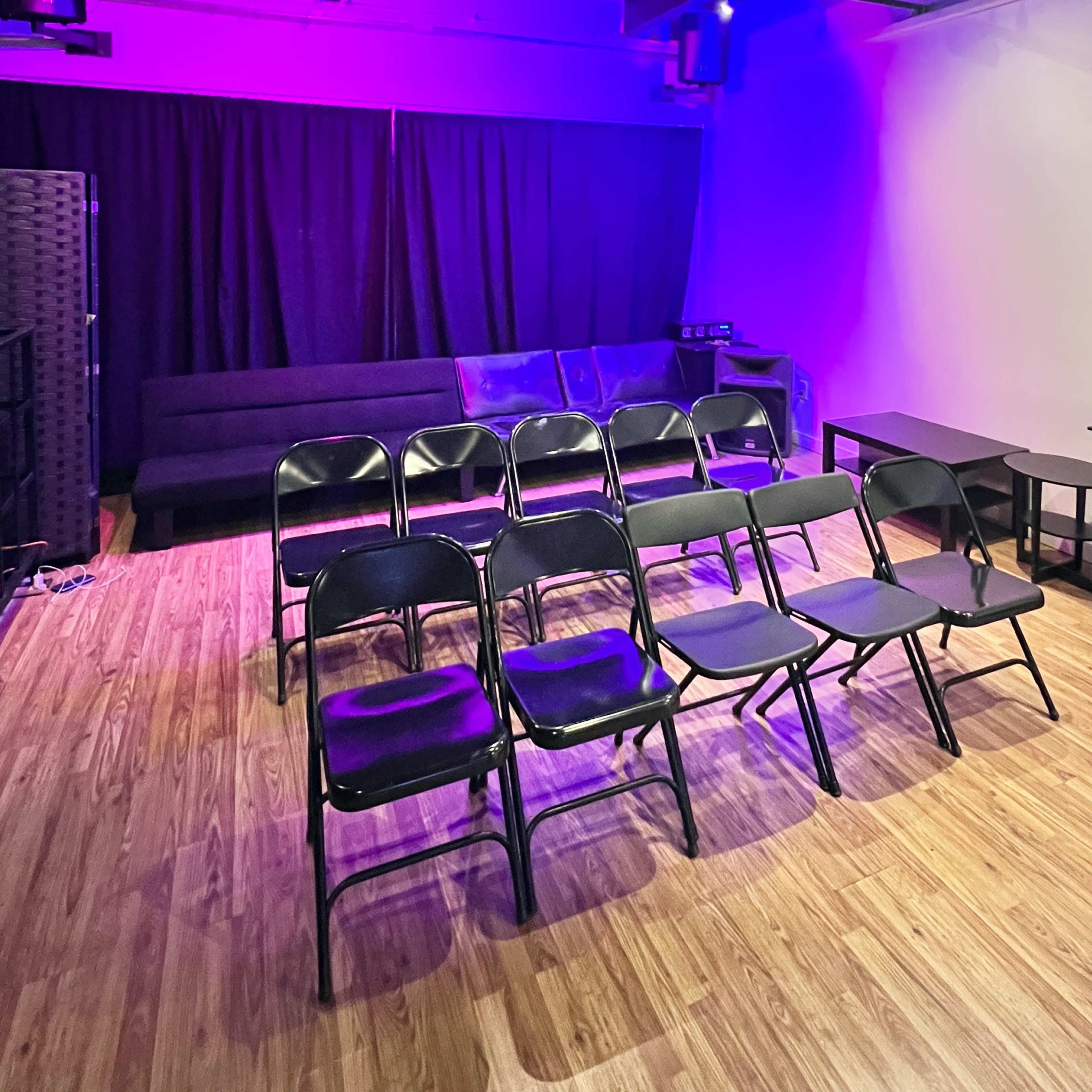 The room features a set of black folding chairs arranged in rows, a dark sofa along one wall, and tables on the other side, with purple lighting illuminating the space.