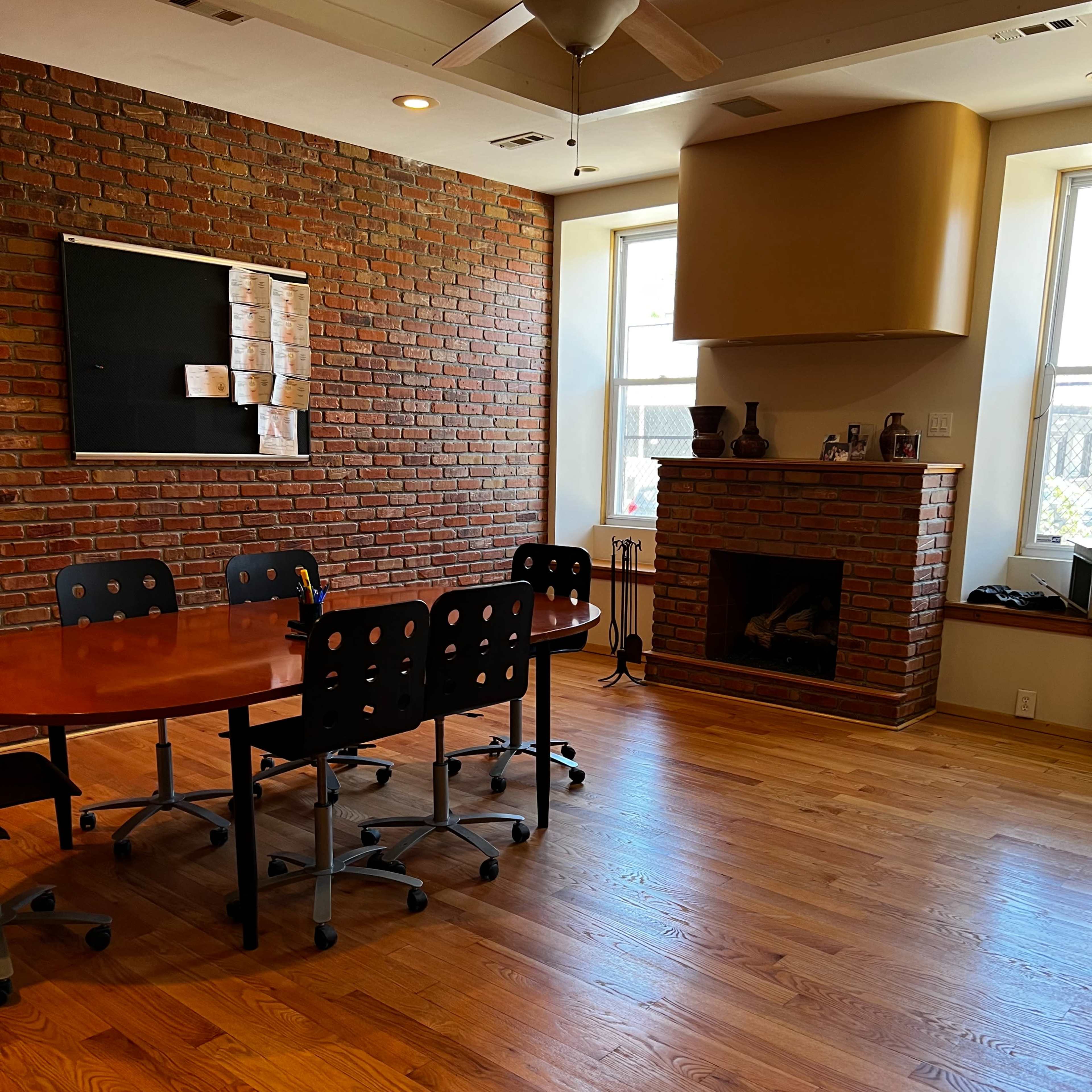 A meeting room with a round wooden table and black rolling chairs, complemented by a brick wall and a fireplace.