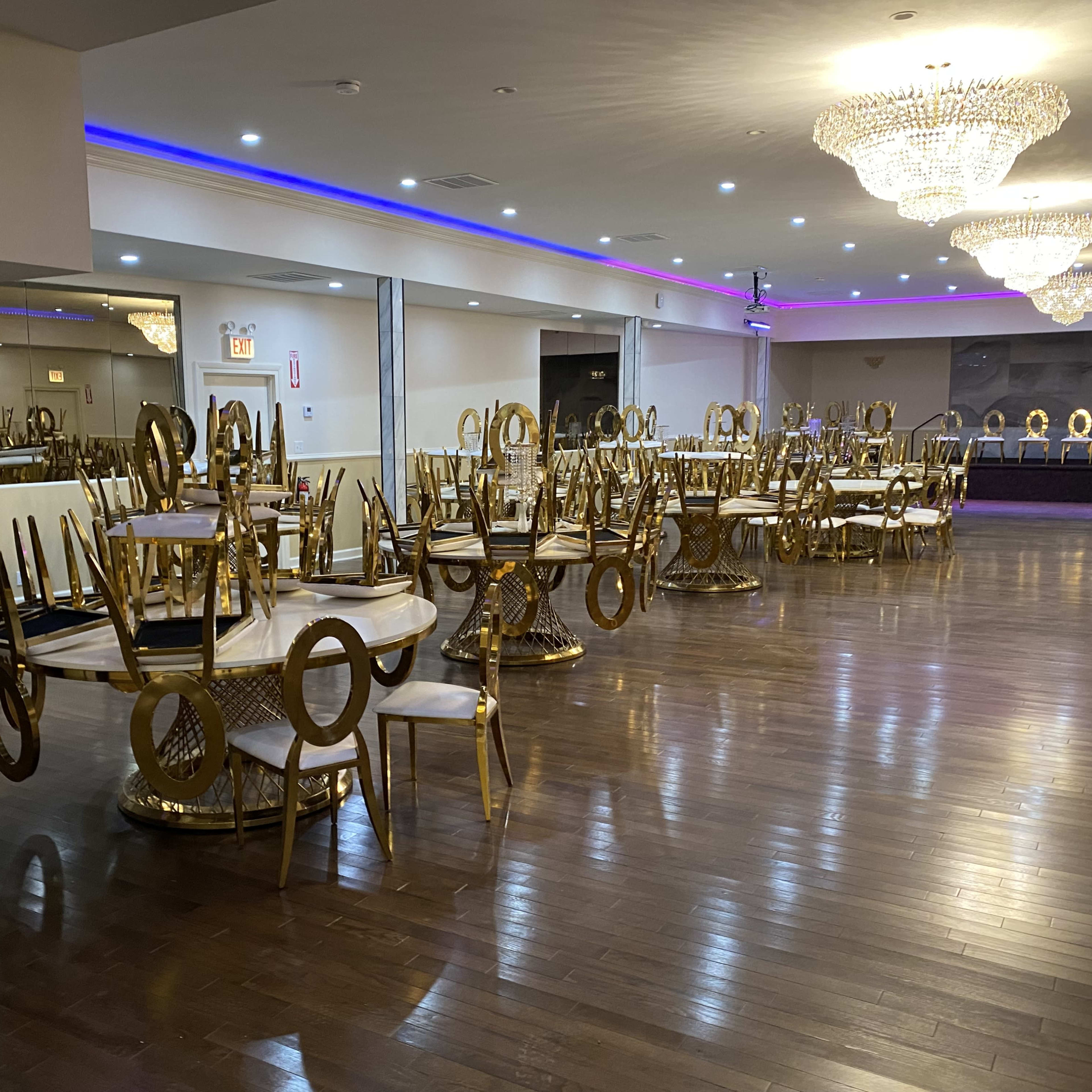 An empty banquet hall with several gold-accented dining tables and chairs stacked upside down, along with decorative chandeliers overhead.
