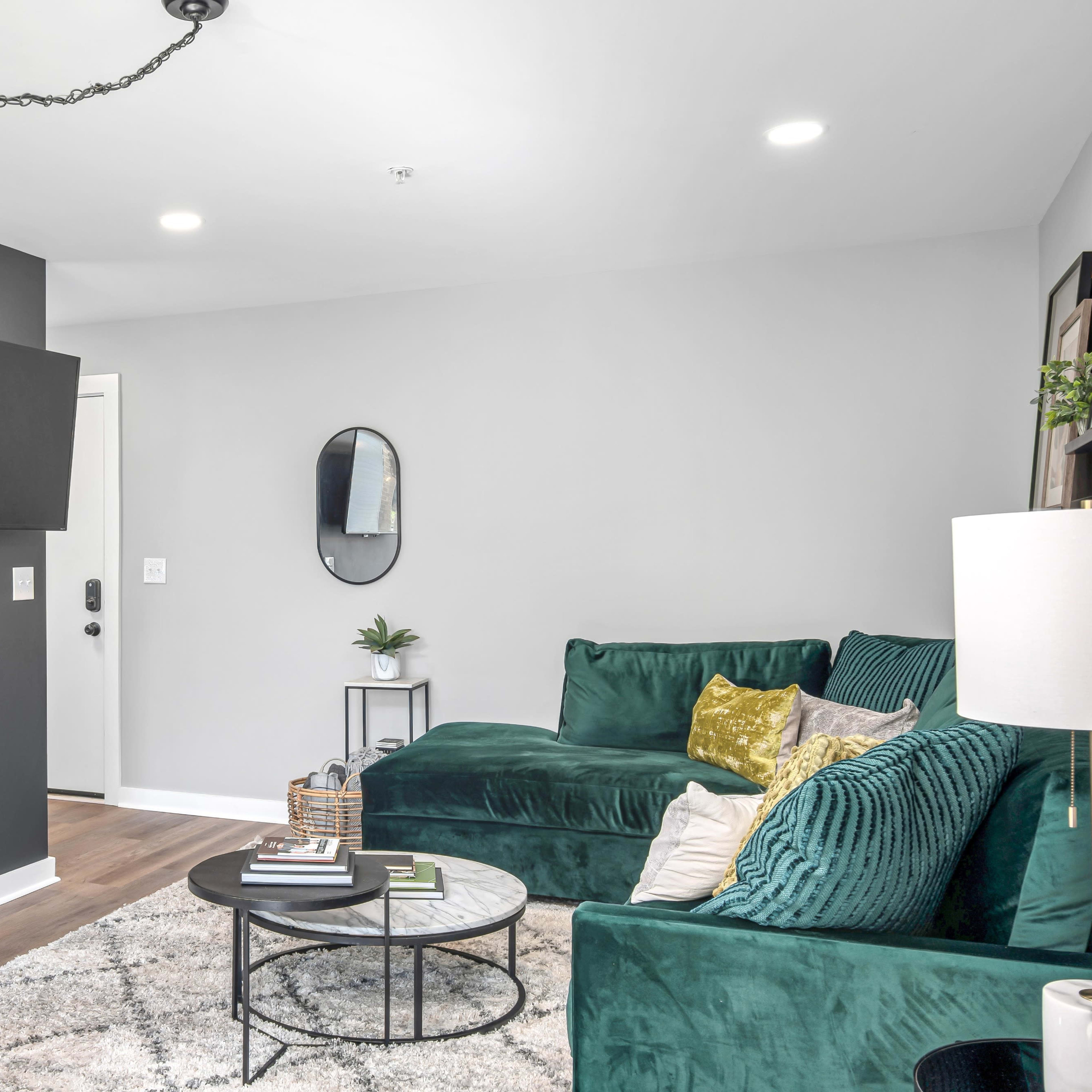 A modern living room featuring a green sectional sofa, a round coffee table on a patterned rug, and a kitchen area in the background with a television mounted on the wall.
