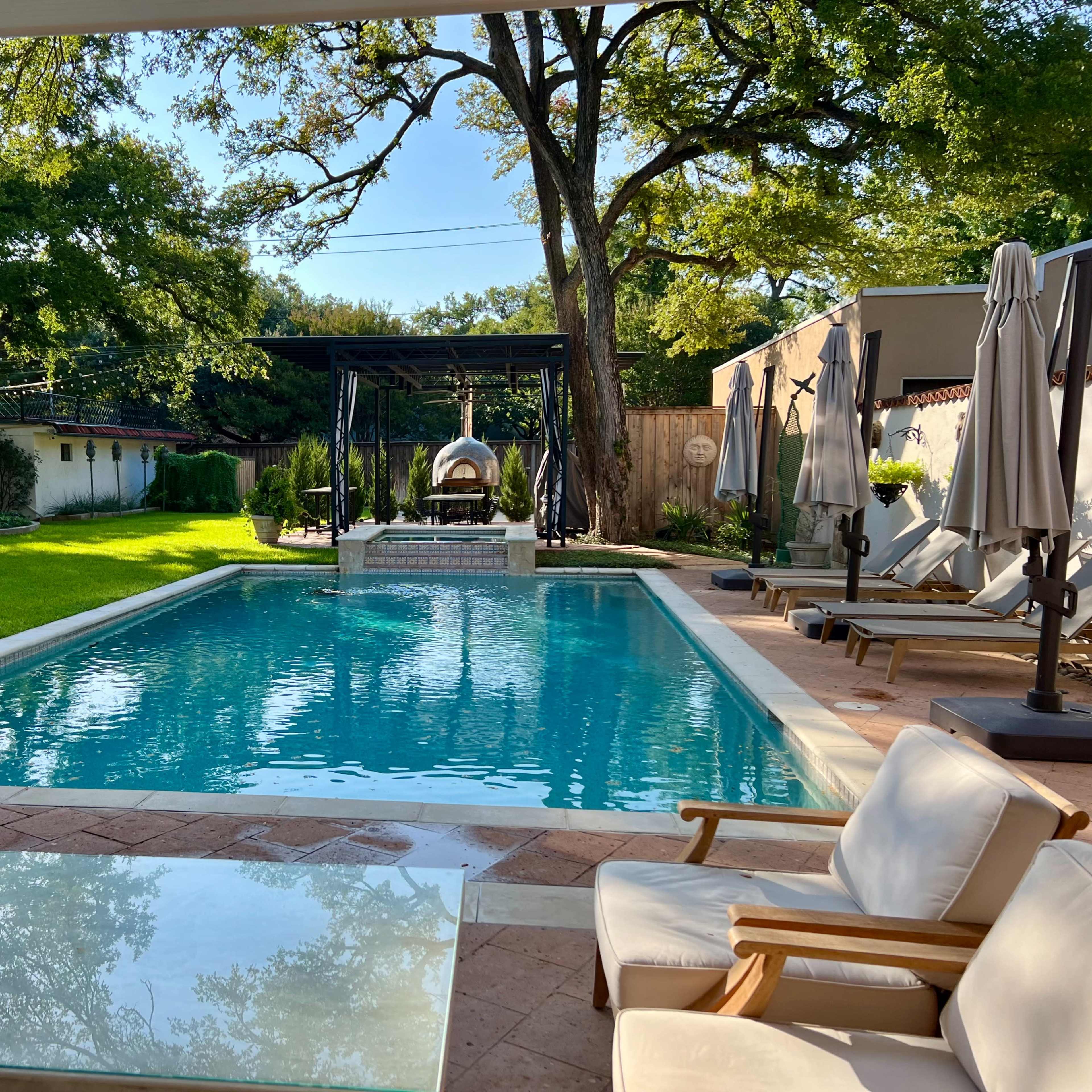 A clear swimming pool surrounded by lounge chairs and greenery, with shaded seating areas and a gazebo in the background.