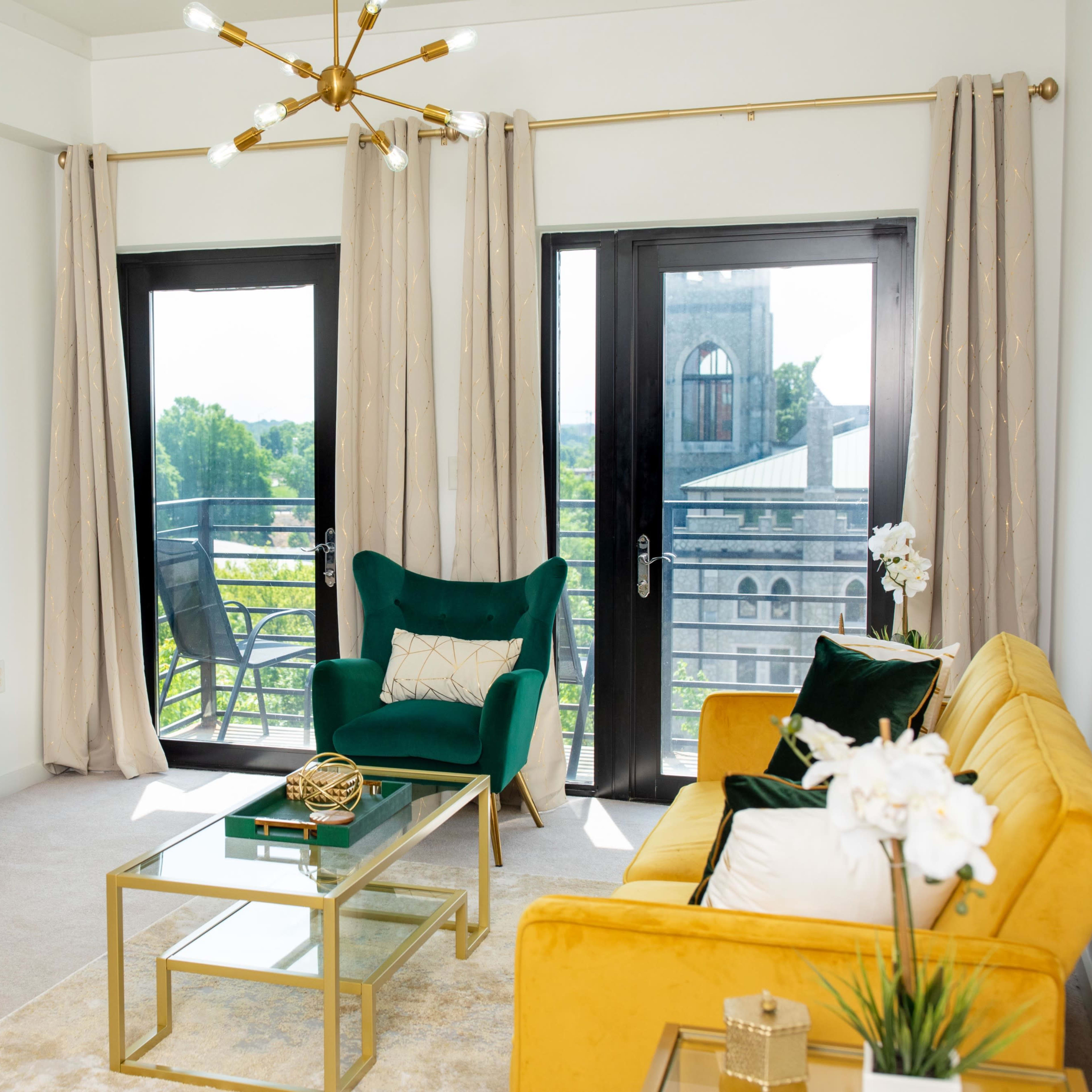 A modern living room featuring a green chair, a yellow sofa, a glass coffee table, and a television on a wooden stand, with large windows overlooking a balcony.