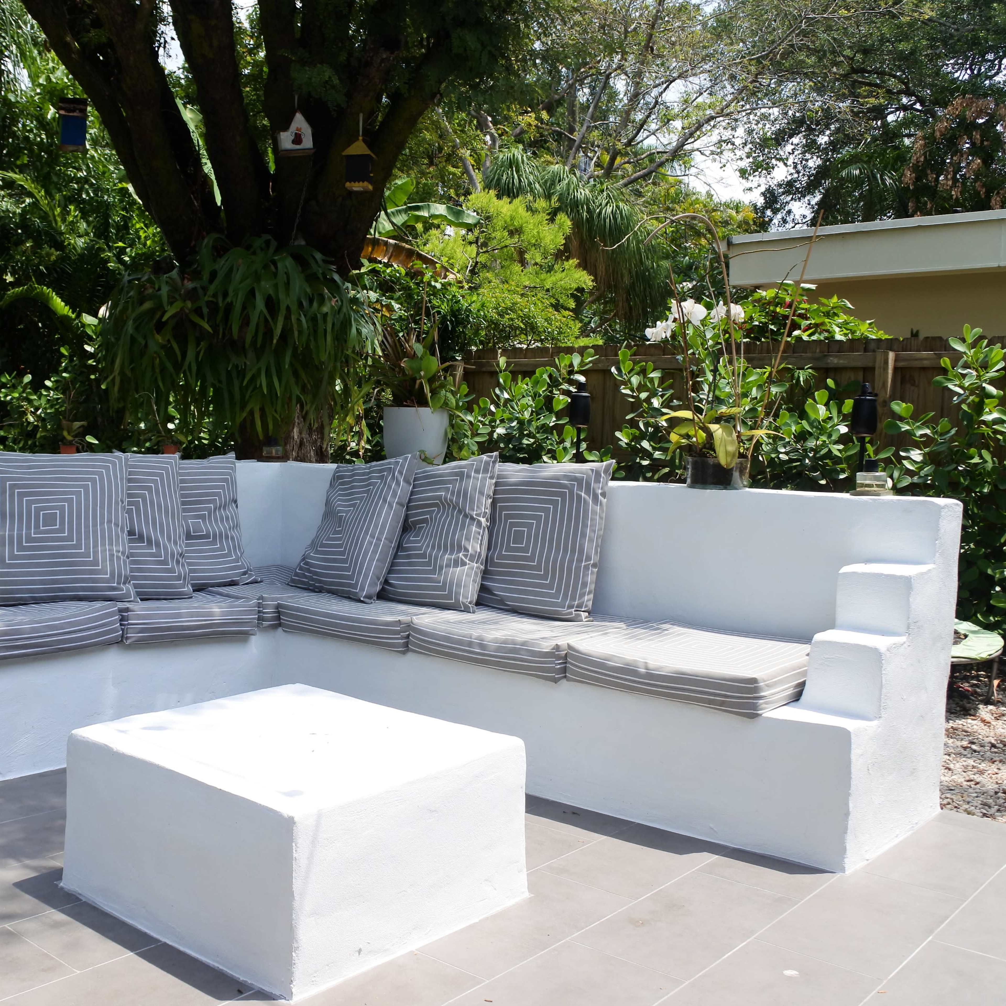 A white outdoor seating area with a long bench covered in striped cushions and a square table, surrounded by greenery and trees.