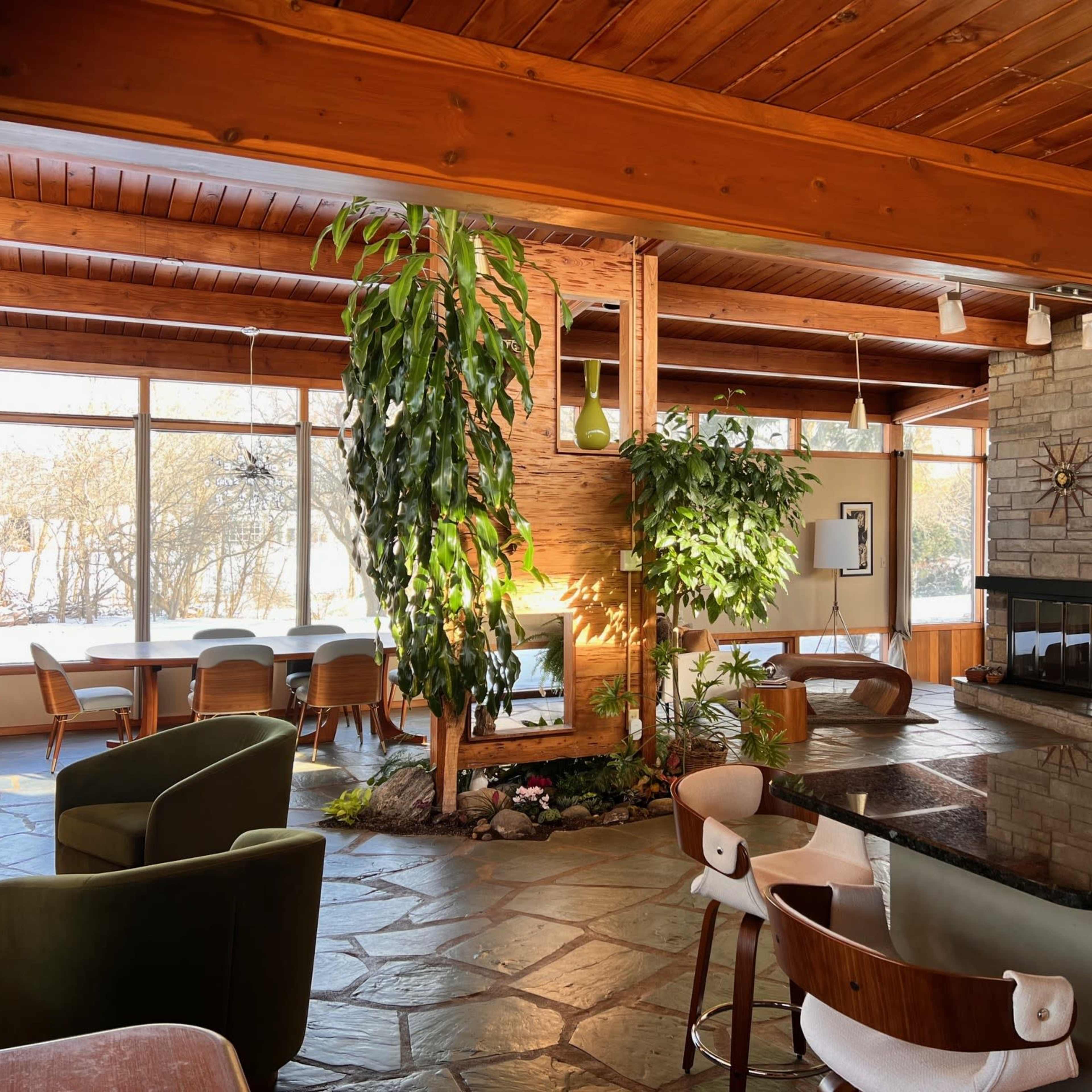 A spacious interior with a stone fireplace, large windows, plants, and a kitchen area featuring a dark countertop.