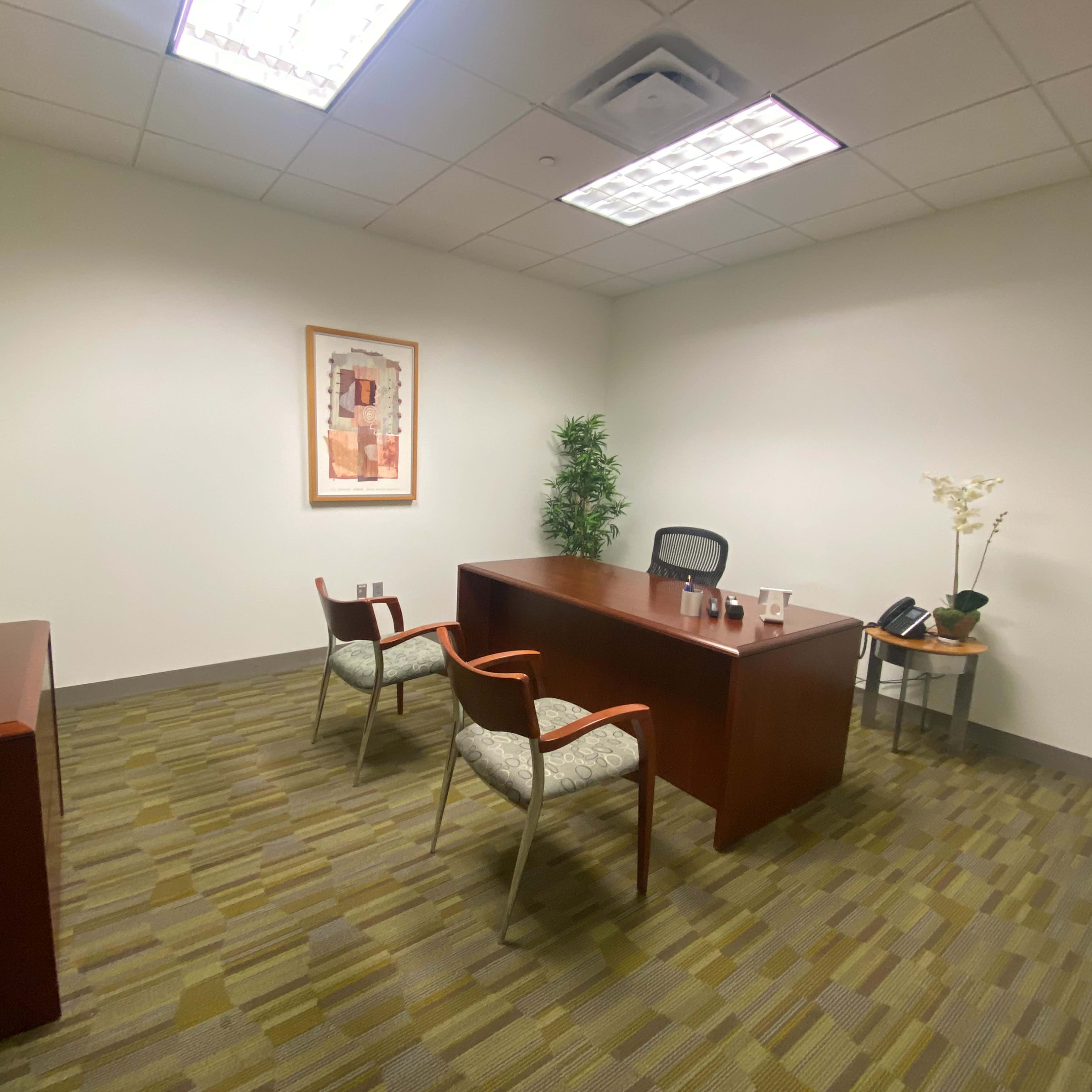 An empty office space features a wooden desk, two chairs, a bookshelf, and a plant against a neutral wall.