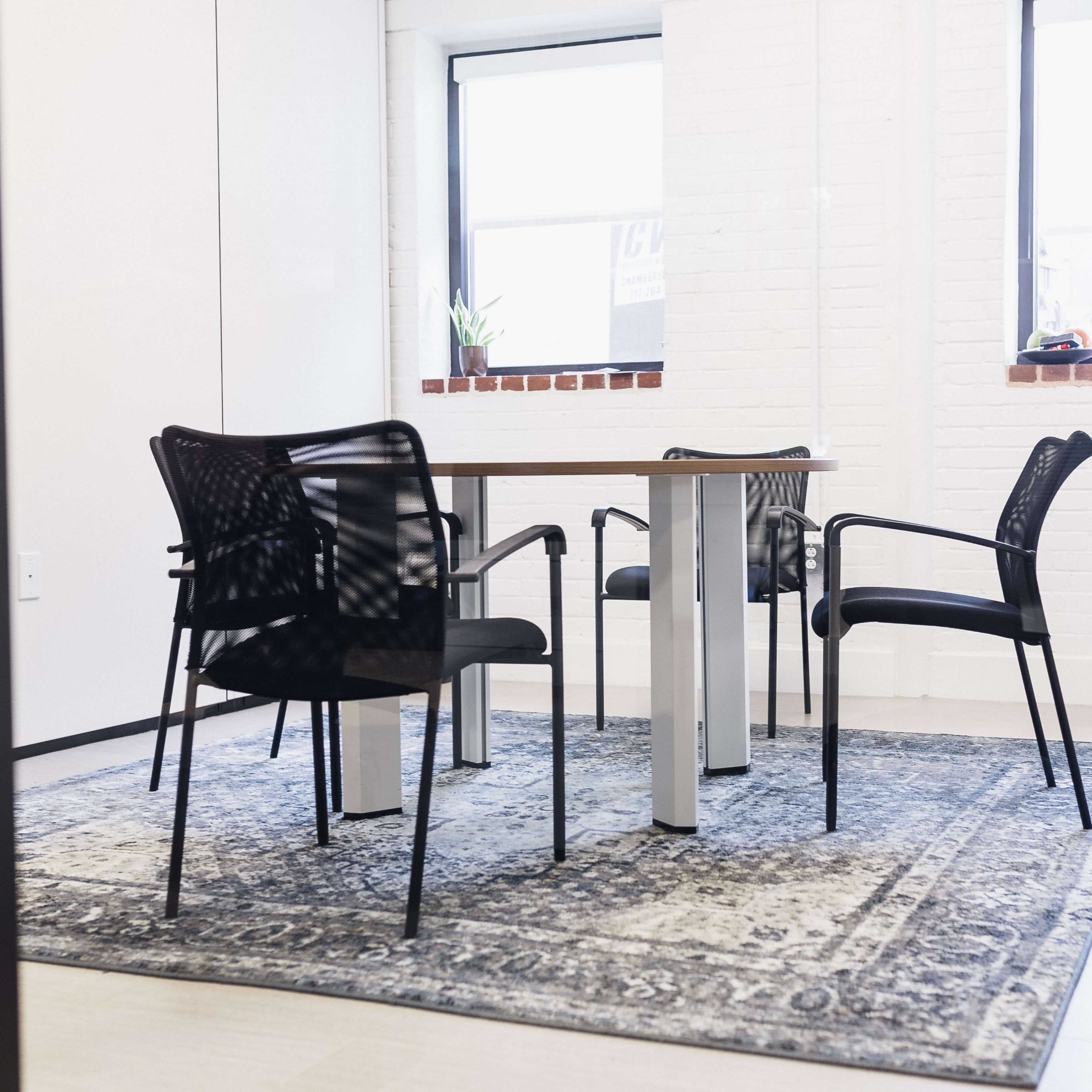 A small meeting room with a wooden table surrounded by four black chairs on a patterned rug, with large windows letting in natural light.