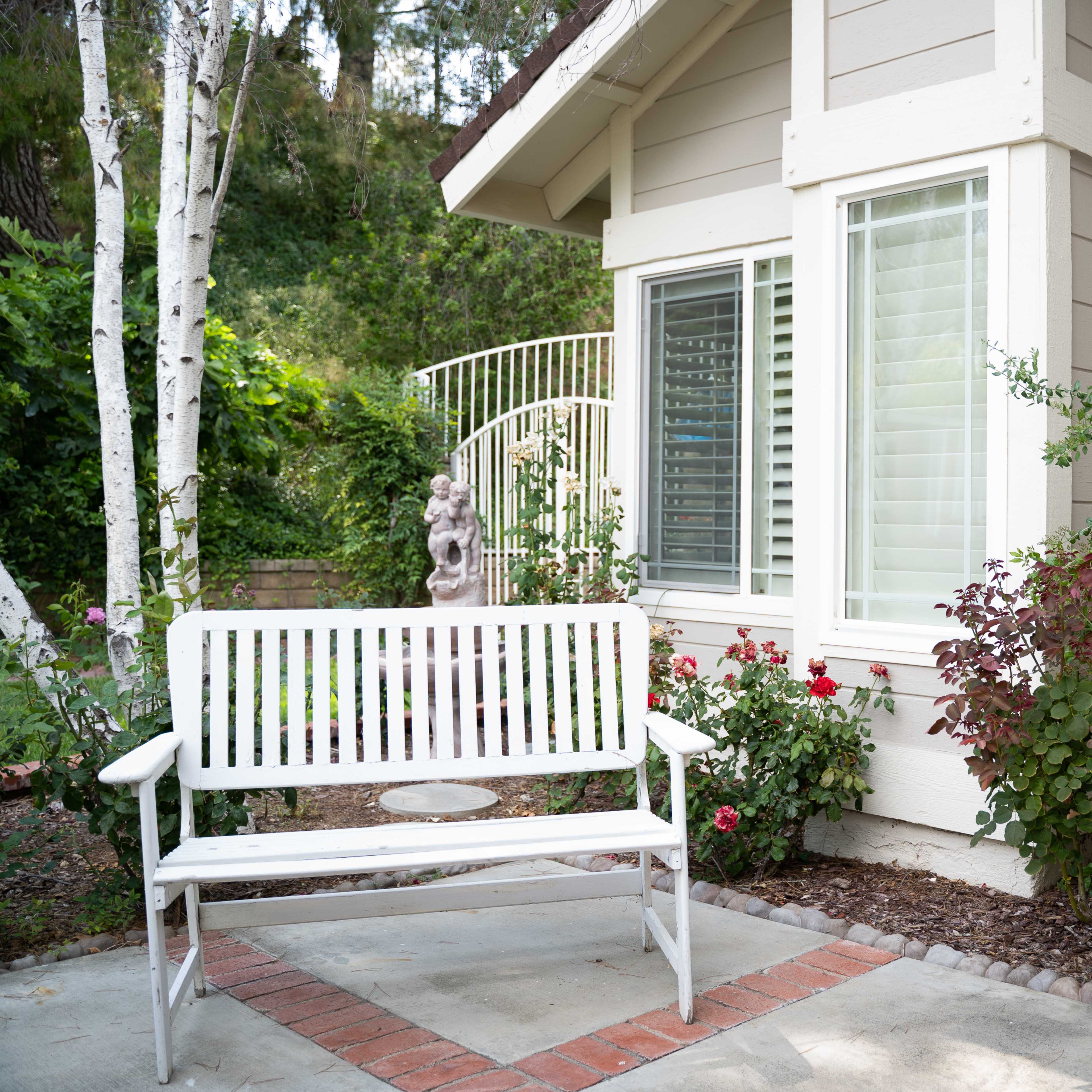 A white bench sits on a patio near a house with green plants and bushes, framed by a white trellis and decorative statue.