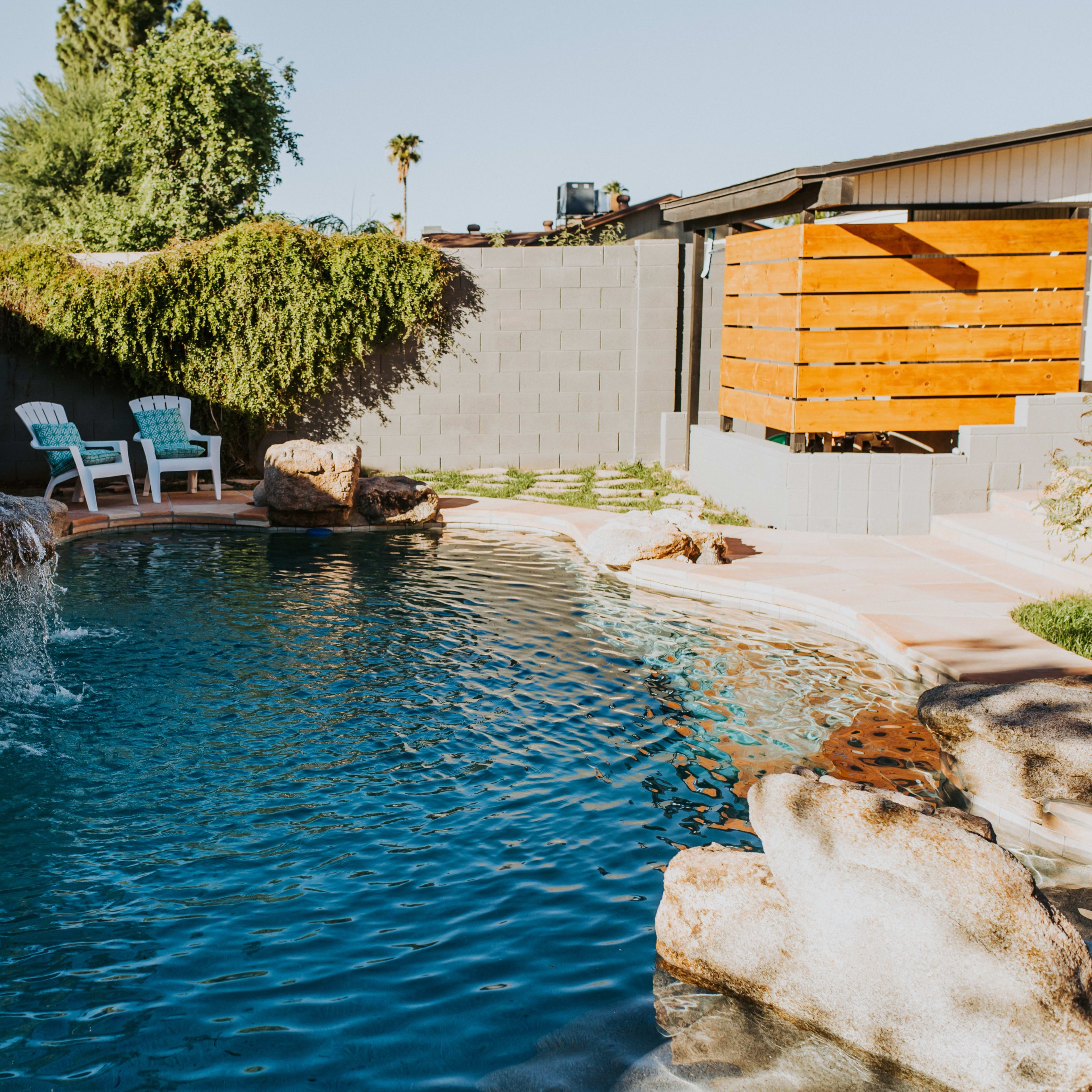 A blue swimming pool with a rock waterfall is surrounded by a landscaped yard featuring two white chairs.