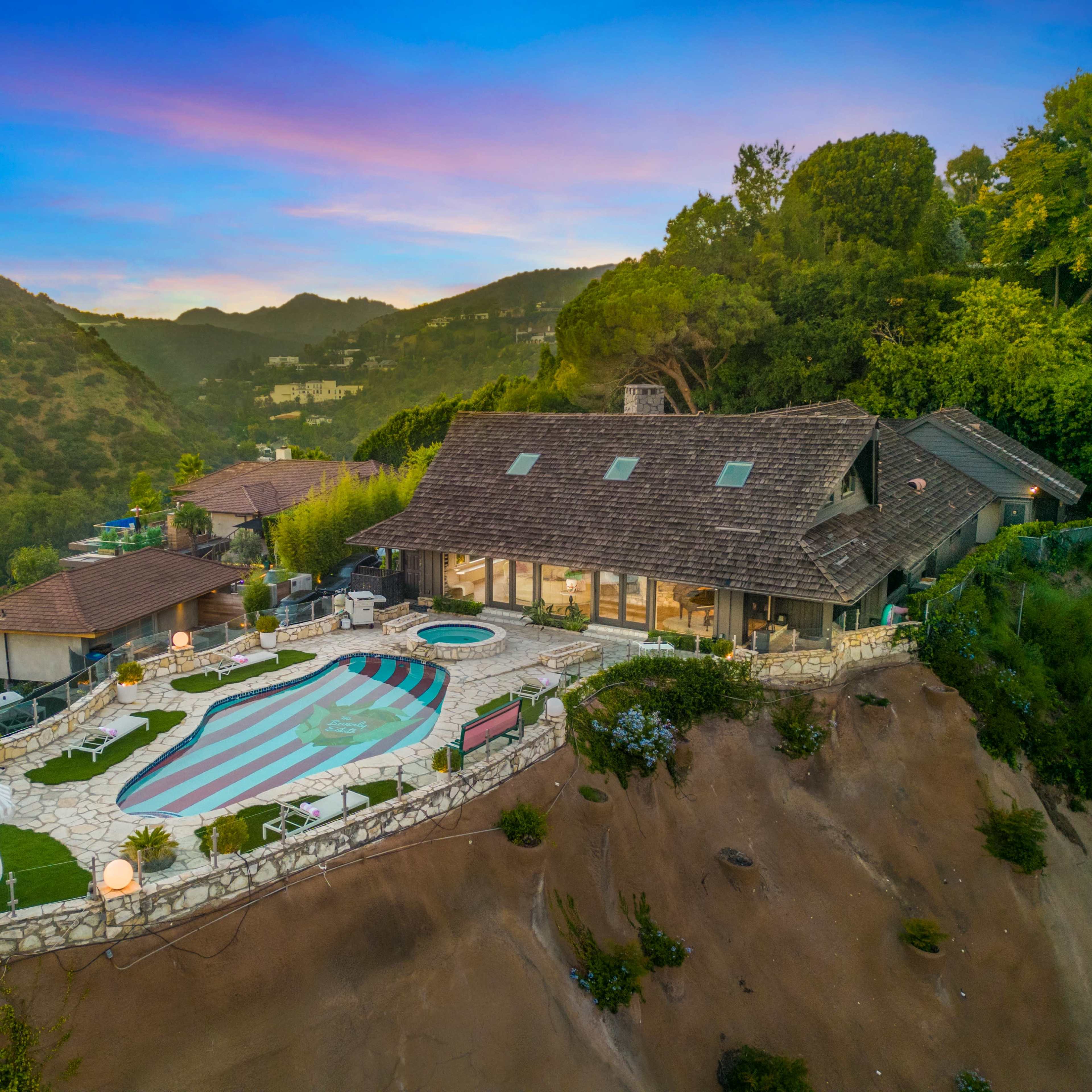 A large, modern house with a swimming pool and landscaped yard, situated on a hillside surrounded by mountains.